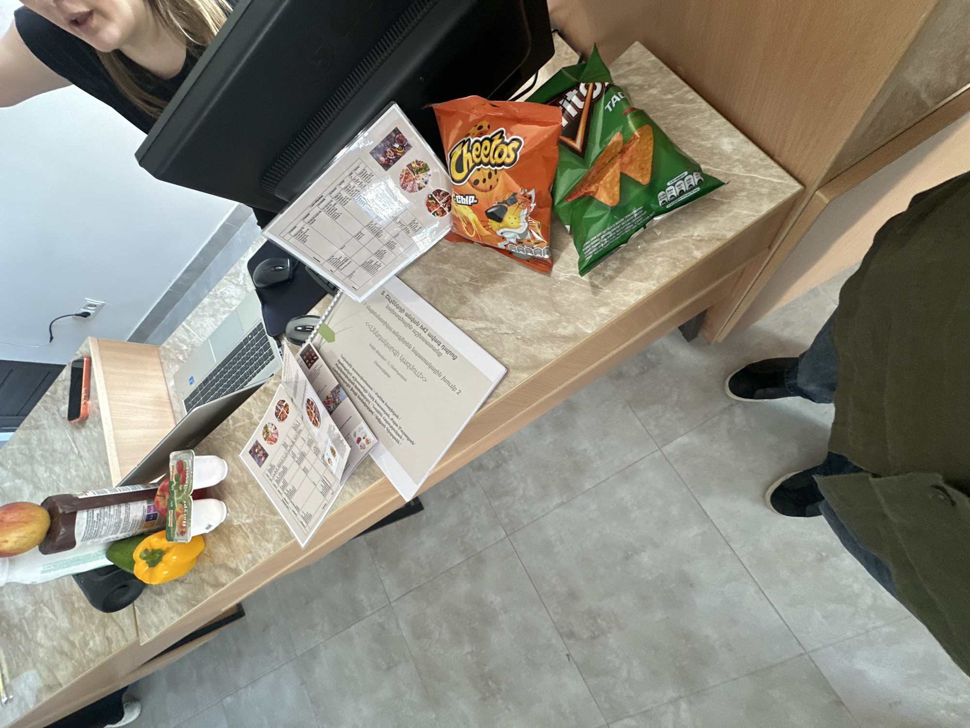 Here is a detailed description of the image:
**Scene:** The scene appears to be a workspace, likely a desk or table area, indoors. The area is cluttered with work-related items and snacks.
**People:** There are parts of two people visible in the image. One person, presumably standing next to the table, has their legs and a portion of a green jacket visible. The second person's head and shoulder is visible at the upper left side of the frame.
**Subjects:** The main subjects are the arrangement of objects on the desk.
**Objects:**
* A computer monitor and a laptop are present.
* Two bags of snacks: one bag of "Cheetos" and one of "Tacos" are visible
* Printed documents, possibly papers or notes, are scattered on the desk. One of the documents has text in a language other than English. The text on the document mentions "Տ. Շևչենկոյի անվան հ42 ավագ դպրոց" (likely a school name) and the topic of "Սննդակարգի կազմում" (potentially "Diet composition").
* A collection of fruits and vegetables including an apple, pepper, and a brown condiment bottle.
* Two white plastic containers.
* A mouse
* A phone
**Actions:** The actions are not immediately evident in the photo. It seems the person was working at the desk, possibly with snacks nearby.
**Interactions:** The arrangement suggests someone was in the process of working and snacking at the desk.
**Environment:** The environment seems to be a working space, probably an office.
**Location:** There is no discernible evidence indicating a specific city or region.