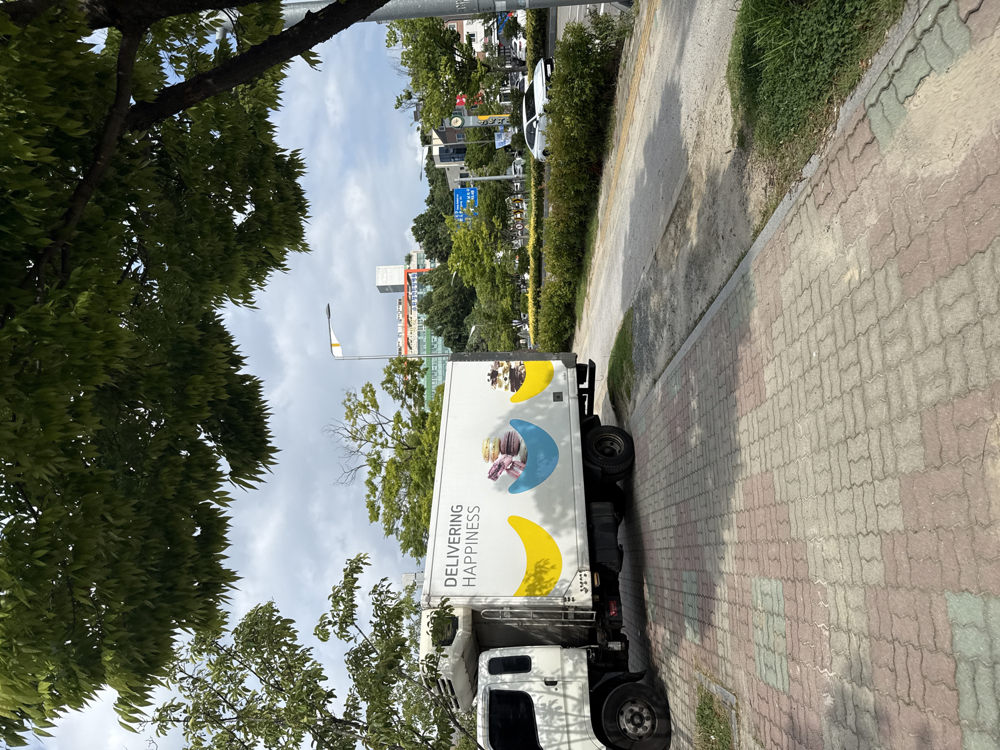 A white delivery truck is parked on a paved walkway in Gwangju, South Korea. The truck has "DELIVERING HAPPINESS" printed on its side, along with graphics of colorful macarons and bananas. The walkway is made of interlocking red and grey bricks, with patches of green grass bordering it. In the background, a street runs alongside a row of green bushes. A white car is visible on the street, as well as several buildings and trees. The sky is overcast with scattered clouds.
