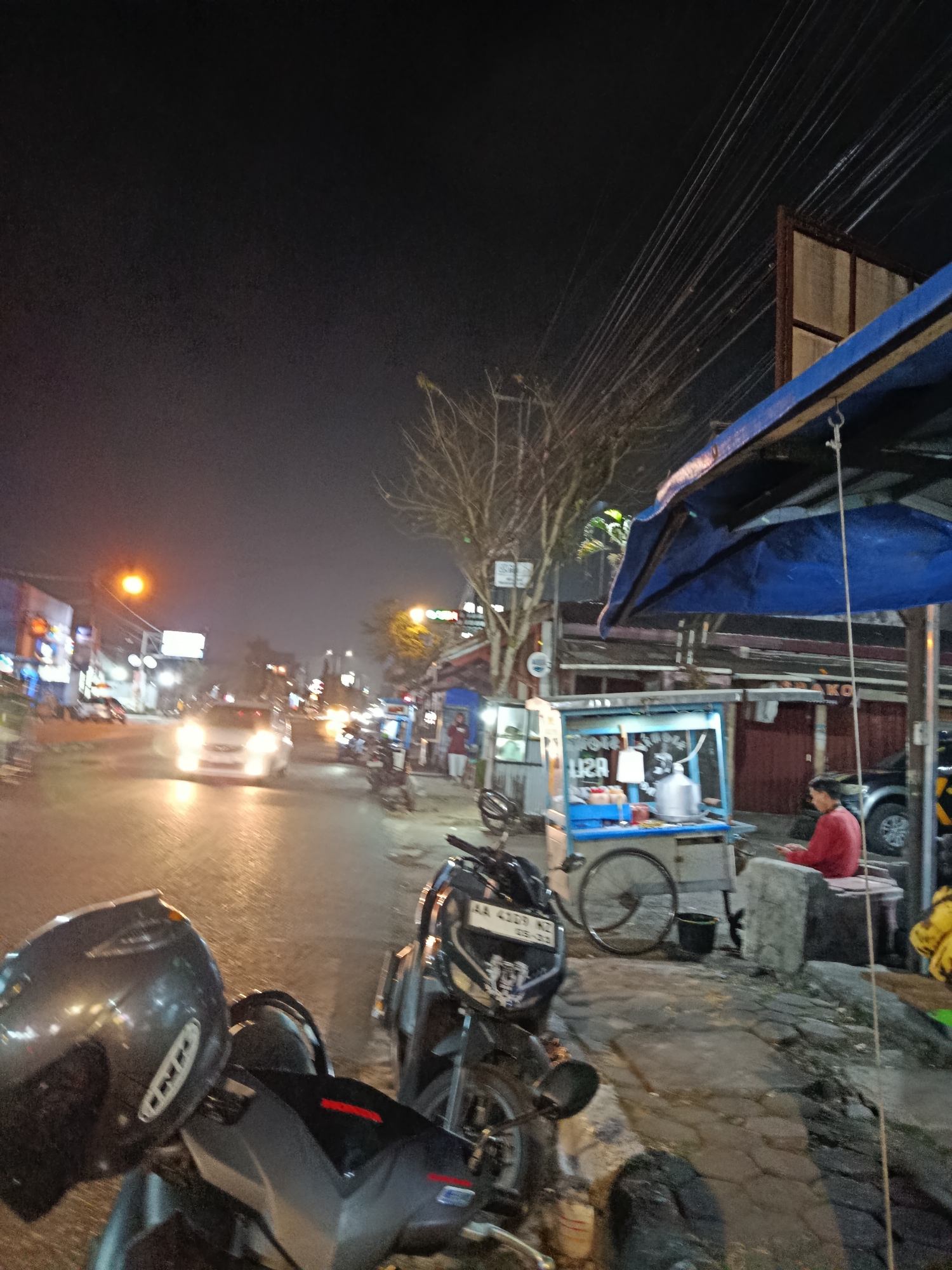 This is a night-time street scene in Mlipak, Indonesia, featuring a road with vehicle traffic and a pedestrian walkway lined with vendors. In the foreground, two motorcycles are parked, one with a dark grey helmet placed on its seat. The street is wet, reflecting the bright headlights of a white car driving towards the camera. On the right side of the street, a person in a red shirt is sitting at a food cart that is lit up. The cart appears to be serving food, with various containers and utensils visible. A blue tarp is stretched over a part of the food cart area. The background shows the typical urban nightscape with streetlights, shop signs, and overhead power lines. Trees with bare branches are also visible. The overall atmosphere is that of a busy, active street market at night.