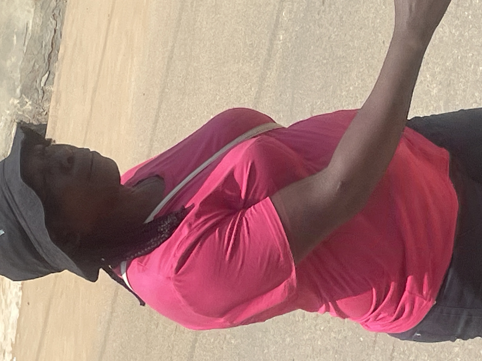 The media file shows a person from the chest down, wearing a bright pink t-shirt and a dark hat. The person has dark skin and their hair is braided and visible. A light-colored strap runs diagonally across the chest of the t-shirt. The person is wearing dark pants. The scene appears to be outdoors with a textured, light-colored ground, possibly sand or pavement. The lighting suggests it is daytime, with bright sunlight casting shadows. The overall impression is of a person standing or walking in an open, possibly urban or recreational area. No text is visible in the image. Given the location context of Abuja, Nigeria, it is likely that this image was taken there. The person's expression is not visible due to the framing of the shot.