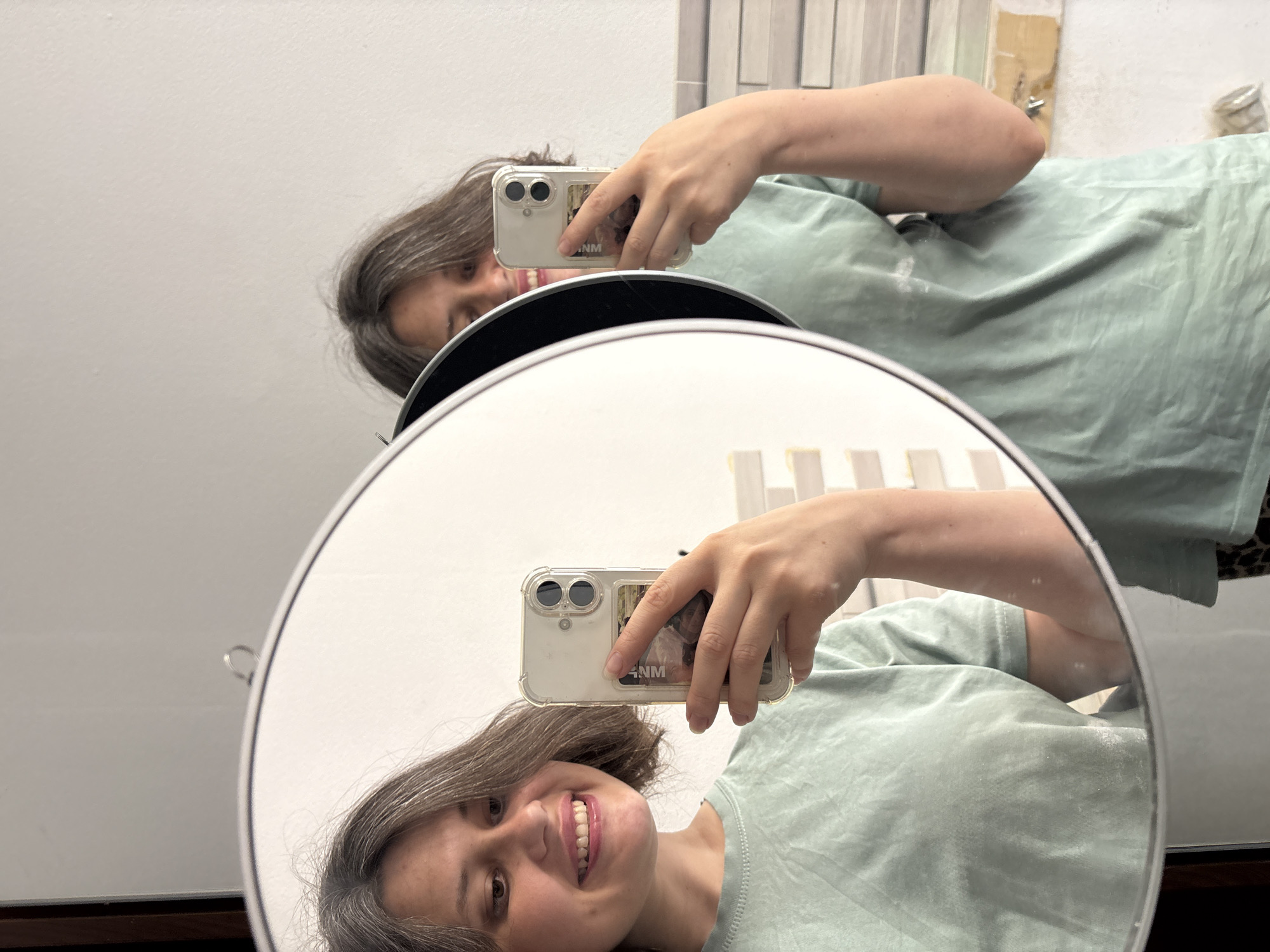 A person is taking a selfie in a round mirror. The person is smiling and holding a smartphone with a clear case in their right hand. The phone case has a sticker on it that reads "NM." The person is wearing a light green t-shirt. The background appears to be a bathroom or dressing room with tiled walls and a wooden countertop. The image does not provide enough information to confirm the location in the city of Chaikovsky, Russia.