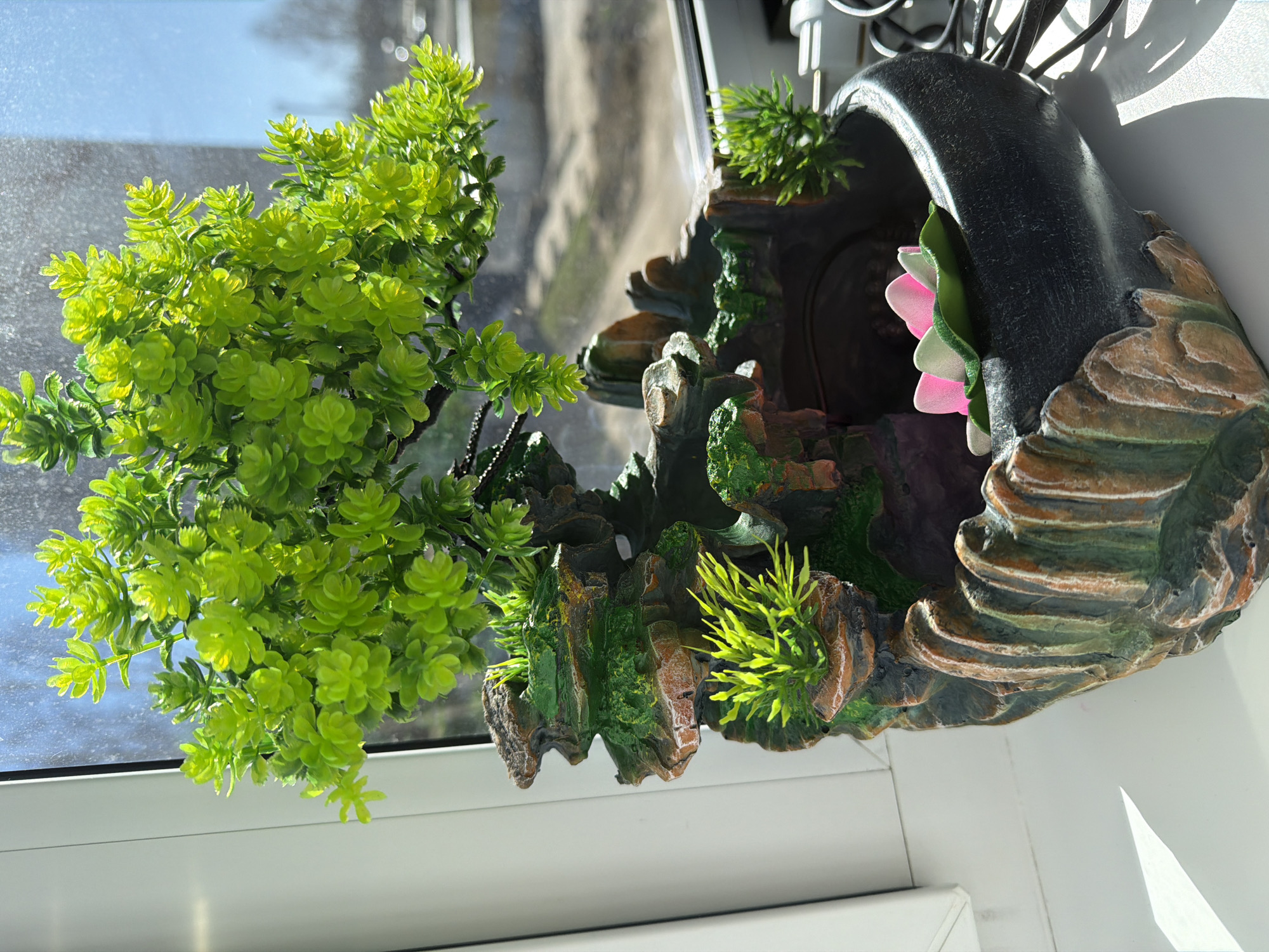 The image shows an indoor scene with a decorative item placed on a white windowsill. The decorative item consists of two parts: a green artificial plant on the left and a stone-like structure with a dark arch on the right. The stone-like structure has a pink artificial flower attached to the side of the arch. The background visible through the window appears to be an urban setting, indistinct and out of focus. The image is taken in daylight conditions. There is no indication of the image being taken in Votkinsk, Russia.