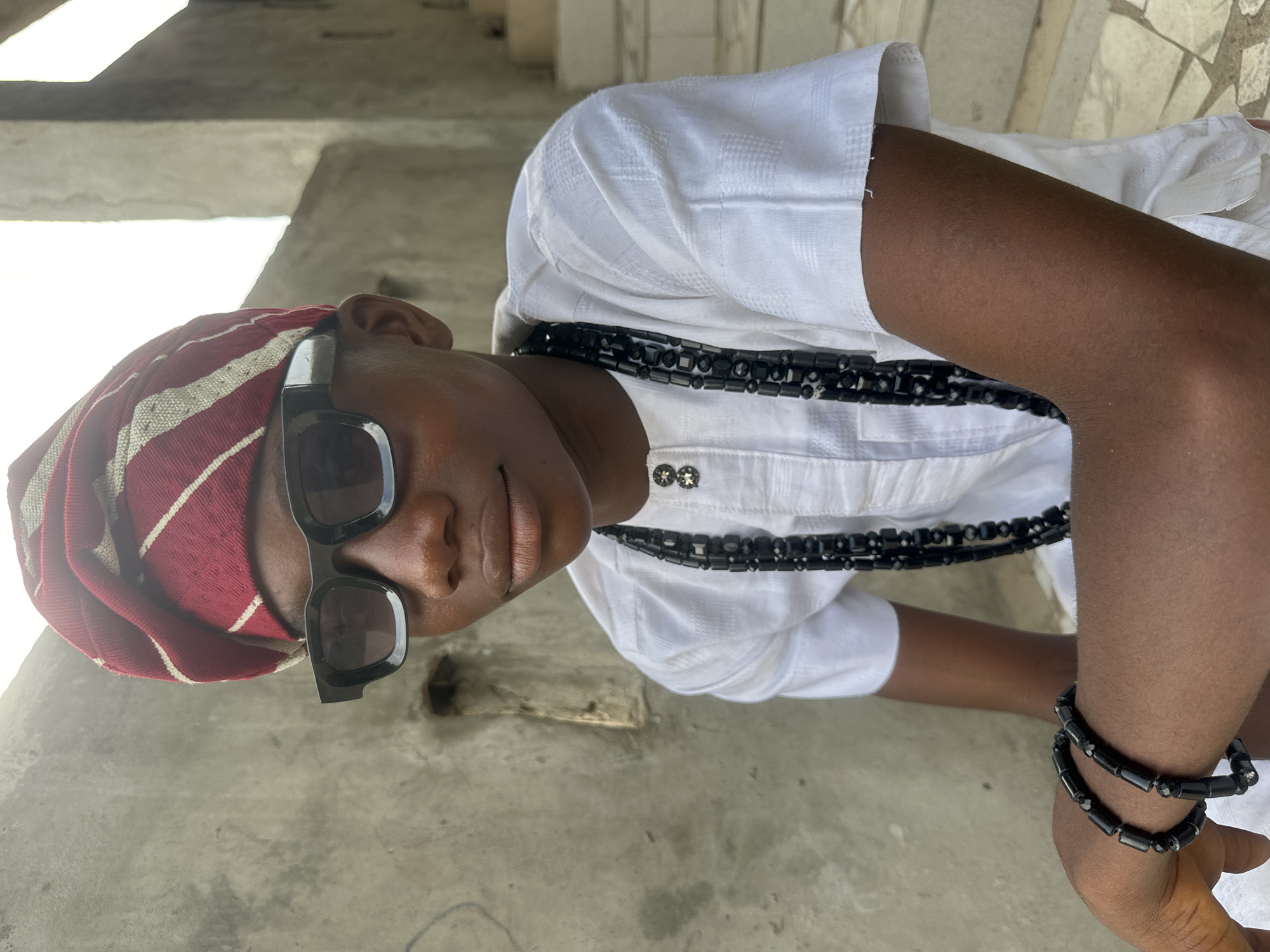 A young Black male, wearing a white shirt, a red and white striped headwrap, dark sunglasses, and black beaded necklaces and bracelets, poses for a photo in front of a weathered concrete wall. He is seated, with his left arm resting on his lap, adorned with two black beaded bracelets. A long strand of black beads hangs around his neck, partially obscuring the top buttons of his shirt. The background consists of rough, textured concrete surfaces and a glimpse of a tiled staircase to the right. The setting appears to be outdoors, possibly in a courtyard or residential area, with the architecture suggesting a location in Badagry, Nigeria. The lighting is bright, casting shadows and highlighting the textures of the clothing and the environment.