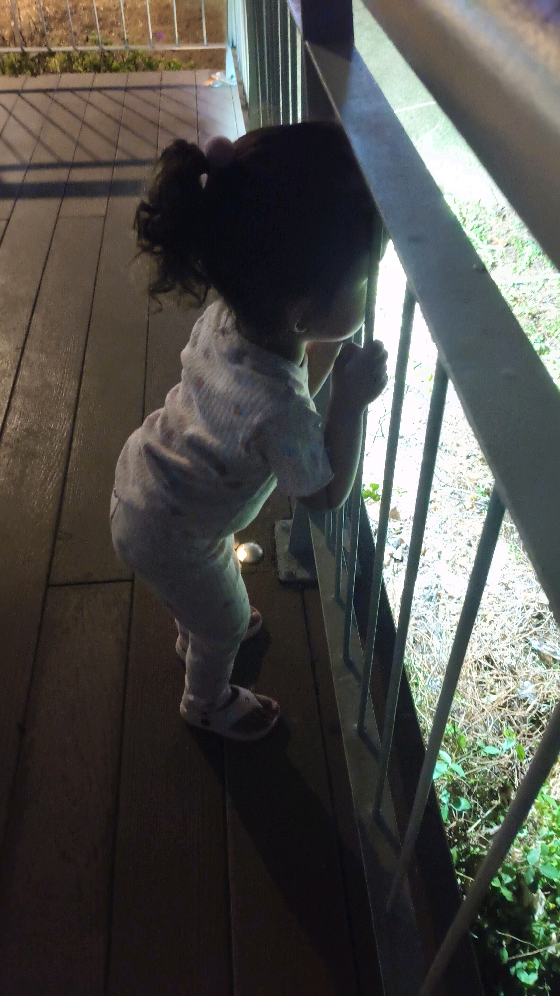 A young child with dark hair in a ponytail, wearing a light-colored pajama set, stands on a wooden deck, leaning over a metal railing to look at the ground below. The child is viewed from behind and to the side. The deck appears to be outdoors, possibly a balcony or patio, with metal railings and a wooden floor. The ground visible through the railings is covered with dry grass and some green foliage, suggesting a natural or overgrown area. The lighting suggests it is either evening or a dimly lit interior space with outdoor lighting. The scene is set on a wooden deck with a railing, with the child's attention directed downwards through the railing. The overall impression is of a child exploring or observing their surroundings from a raised vantage point. The image does not contain information that would allow for specific identification of the location in West Jakarta, Indonesia.