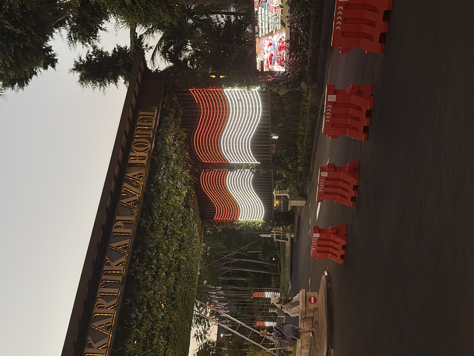 The image depicts the entrance to the Kartika Plaza Hotel in Kuta, Indonesia, at dusk. A large, illuminated sign spelling "KARTIKA PLAZA HOTEL" is prominently displayed above the entrance, with the letters lit in gold. The entrance archway is covered in lush green foliage. Two large, waving Indonesian flags, rendered in red and white neon lights, are mounted on the right side of the archway. On the left side, a row of national flags of various countries is visible, accompanied by a statue of a lion.
In the foreground, there is a paved road with several bright orange plastic barriers placed across it. Two individuals are visible near the entrance on the left: one person is standing on a raised platform next to the lion statue, and another is walking away from the camera towards the archway. A digital billboard displaying an advertisement is visible on the right side of the entrance. The sky above is a pale, overcast hue, suggesting the end of the day. The overall scene portrays the entrance to a resort or hotel complex, likely a busy area given the presence of multiple flags and the advertising display.