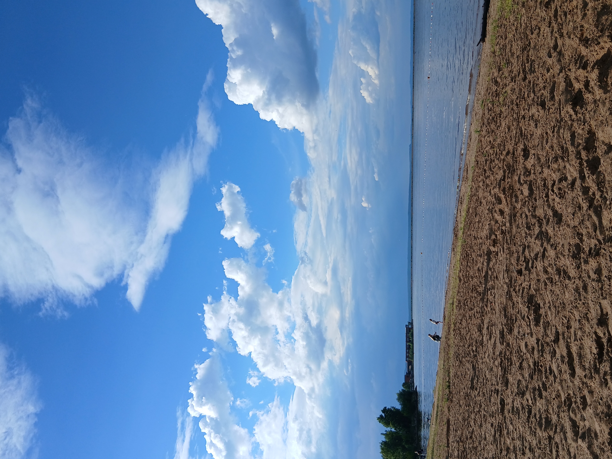 The image depicts a sandy beach on the shore of the Kama River in Chaikovsky, Russia. The sky is a vibrant blue, filled with fluffy white cumulus clouds. The river is calm and reflects the sky. In the distance, a line of buoys marks a swimming area. On the shore, a few people are visible, including two children playing near the water's edge, and a person walking their dog. Behind the beach, there is a line of trees and buildings, suggesting the proximity of a populated area. The overall scene is one of a peaceful summer day by the water.