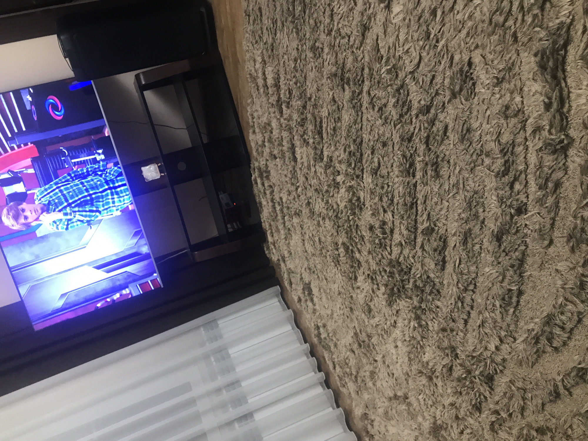 The image shows an interior space, with a focus on the television and nearby furnishings.
A television is displaying a program, featuring a person wearing a blue and white checkered shirt. To the right of the television is a black shelving unit. A set of white curtains are visible below the television.
To the right, the image shows a textured, light brown rug or carpet. The image does not include information to conclude the location in Yerevan, Armenia.