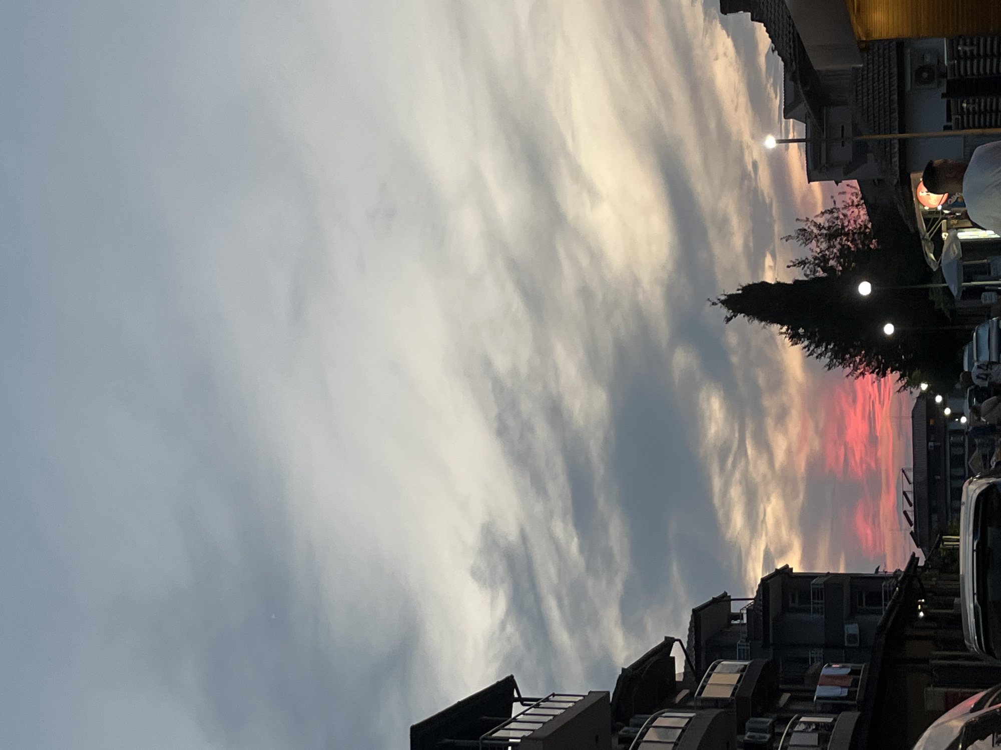 The image captures a street scene in Lozenets, Bulgaria, during sunset. The sky is filled with dramatic clouds, transitioning from a pale blue at the top to vibrant pinks and oranges near the horizon, indicating the sun has recently set. On the left, a multi-story building with balconies and visible air conditioning units is prominent. Clothes are hanging to dry on one of the balconies. In the foreground, the tops of two cars are visible. The street is lined with buildings and streetlights. A tall, dark evergreen tree stands out against the colorful sky. A few people are present, including a man in a white t-shirt walking away from the camera, and other figures further down the street. The overall atmosphere suggests a residential or commercial area in a Bulgarian town at dusk.
