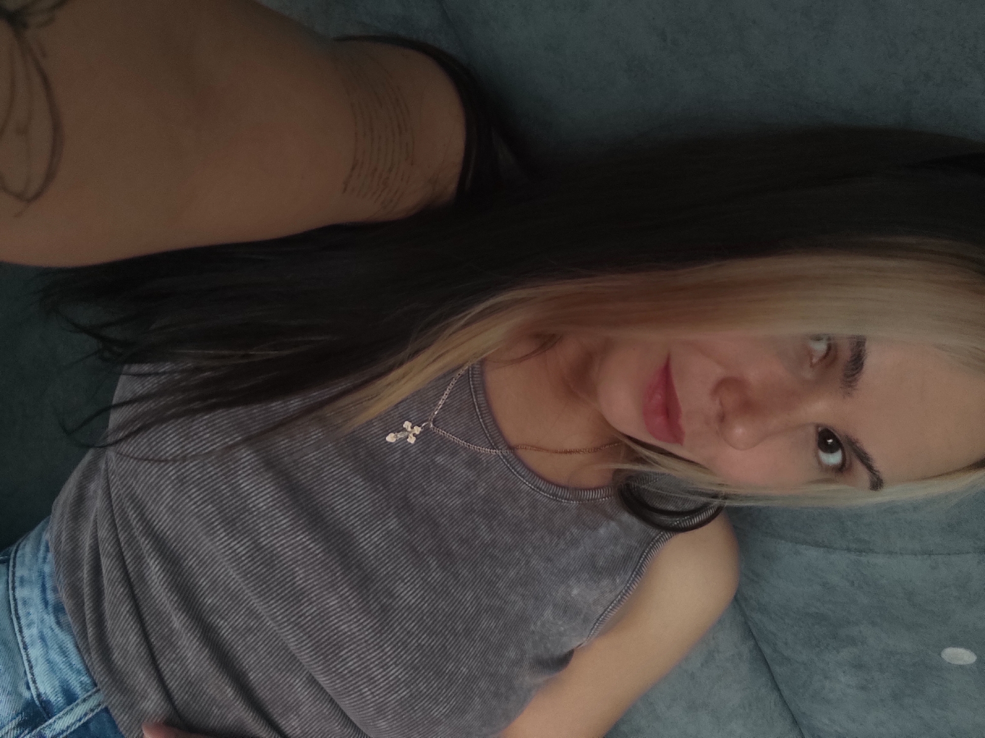 A young woman is lying on a blue-grey sofa or bed. She is wearing a grey tank top and light blue jeans. She has blonde streaks in her otherwise dark hair and wears a necklace with a cross pendant. The image is taken from a high angle, close to the woman's face. Tattoos are visible on her arm. There is no evidence to suggest the location of the photo is Ольховка, Russia.