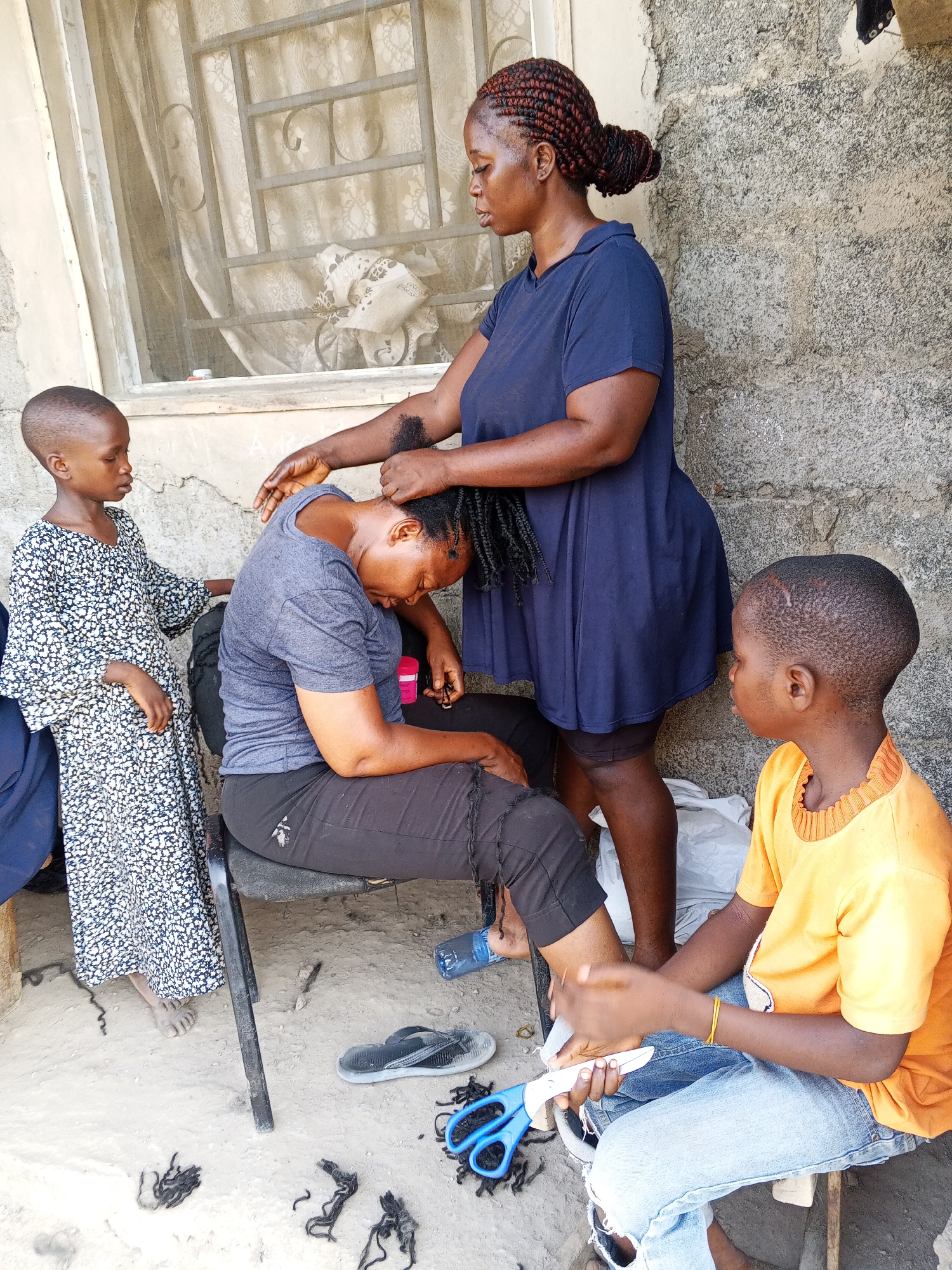 Outside a concrete building in Unknown, Unknown, a woman in a blue dress stands and braids the hair of another woman seated on a black chair. The seated woman is wearing a grey t-shirt and black pants. A young child in a floral dress stands to the left of the braiding women, looking on. Another young child in a yellow shirt and jeans sits on the ground to the right, cutting something with scissors. On the ground are discarded hair pieces, sandals, and other items. A window with a screen is behind the standing woman.