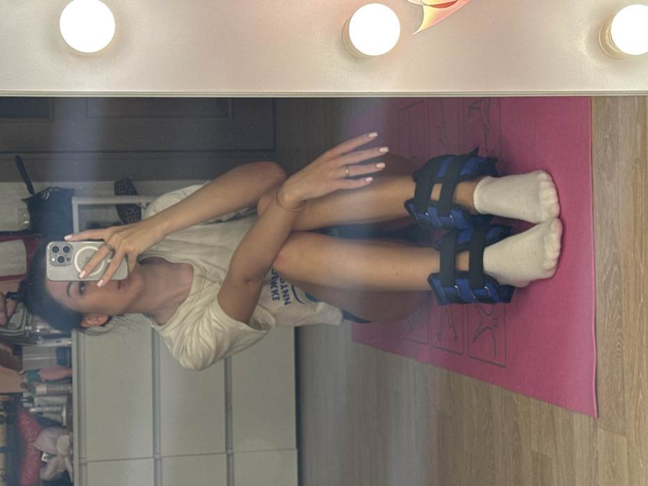 The image captures a mirror selfie, presented sideways, reflecting a person engaged in a fitness-related activity.
The primary subject is a person, likely female, lying on their back on a pink yoga mat. Their body is positioned with knees bent and drawn towards the chest, and they are wearing blue and black ankle weights strapped around both ankles, over white socks. The person is dressed in a light-colored (possibly off-white or light gray) short-sleeved t-shirt and dark shorts. Their dark hair appears to be pulled back.
The person is holding a silver smartphone with a white circular MagSafe-compatible accessory on its back, capturing their reflection in what appears to be a large mirror. Their face is visible in profile, looking towards the camera. Notable accessories include a thin ring on the left ring finger and a delicate, thin bracelet on the right wrist. Their fingernails are painted with light pink polish.
The pink yoga mat has outlines of figures demonstrating various exercises or poses printed on its surface, some of which are partially visible beneath the person's body and near their feet. The mat is placed on a light-colored wooden floor.
The reflection in the mirror reveals more details of the setting. Along the top edge of the mirror (which appears as the right edge in the current sideways orientation), several bright, round vanity-style lights are visible, illuminating the scene. In the background of the reflection, to the left of the person (which would be to their right in a correctly oriented view), there is a light-colored wardrobe or set of cabinets with multiple doors or panels. To the right of the person's reflection (appearing on the left in the current image), there are shelves filled with various items, possibly bottles, jars, and other personal effects, along with a dark bag or container.
Partial text is visible on the person's t-shirt, appearing to be "SNN UMKKE NNTS". The overall impression is of someone taking a break or preparing for an exercise routine in a personal indoor space, possibly a bedroom or home gym.