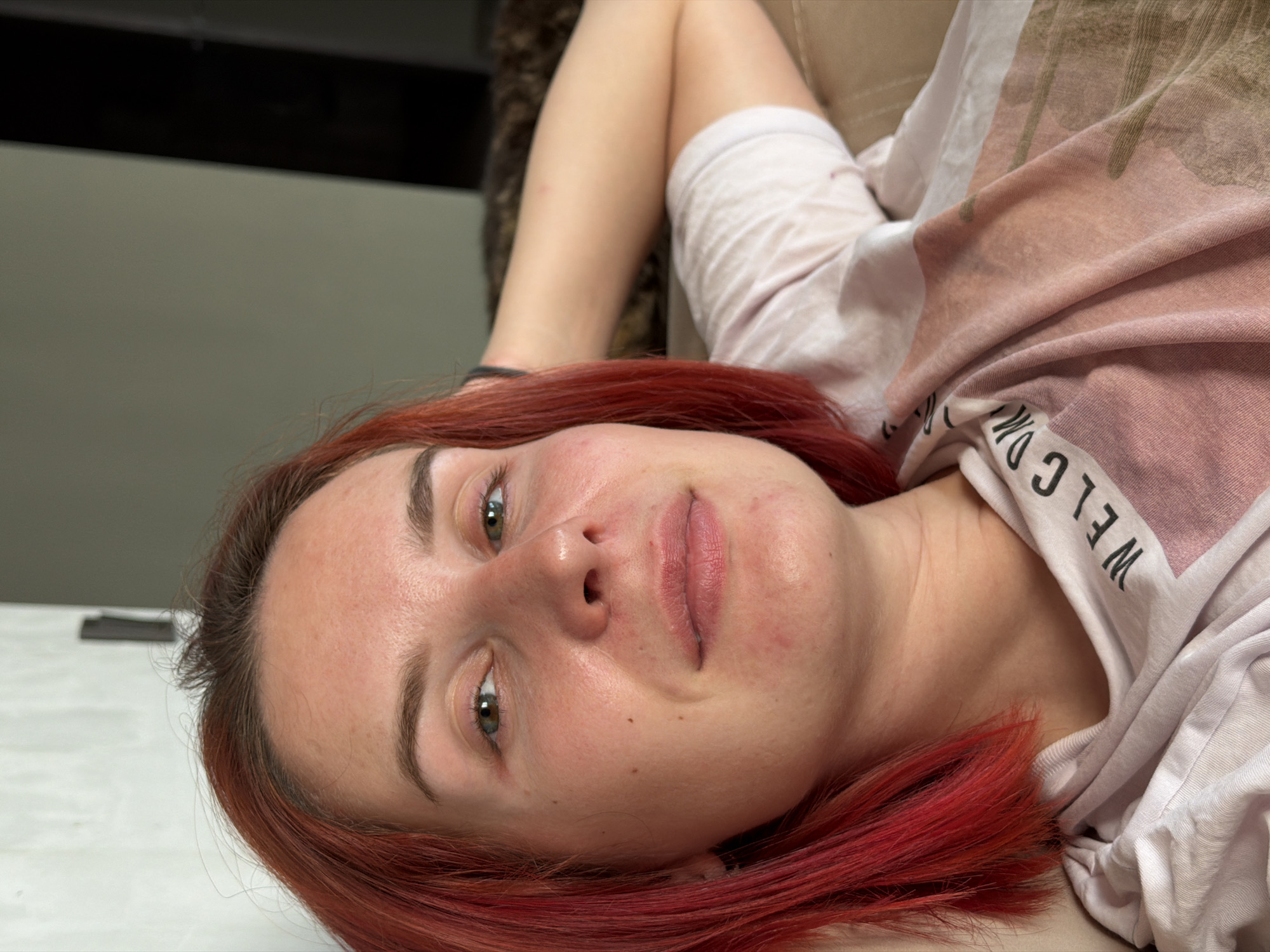 A woman with red hair is lying down. She has fair skin and green eyes. She is wearing a white t-shirt with text on it. The background includes a wall and a portion of what appears to be a couch or similar furniture. There is no information to establish the location as Chaikovsky, Russia.