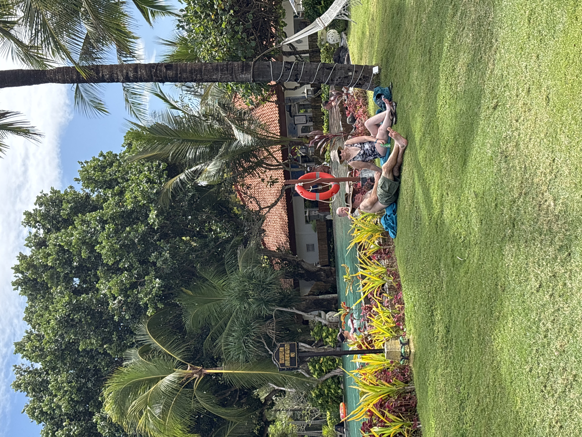 In Kuta Selatan, Indonesia, a tranquil scene unfolds by a resort pool, bathed in the warmth of a sunny day. Two individuals, a man and a woman, recline on the lush green lawn, their bodies angled towards the vibrant turquoise water. The man, clad in olive green shorts and a hat, is partially obscured by a blue towel, his legs outstretched. The woman, dressed in a patterned swimsuit and a sun hat, sits with her legs tucked beneath her, a patterned bag resting beside her.
Behind them, the pool area is a hub of activity. A bright orange life preserver hangs on a pole, a silent guardian by the water's edge. Lush, tropical foliage frames the pool, with vibrant yellow and red plants lining its border. In the distance, a resort building with a distinctive terracotta roof stands as a testament to the region's architectural style. Palm trees, their fronds swaying gently, cast dappled shadows across the scene, adding to the tropical ambiance. A white hammock, strung between two palm trees, offers a glimpse of relaxation, while a coconut tree with a spiraling trunk adds a unique touch to the landscape. The sky above is a clear blue, dotted with a few fluffy white clouds.