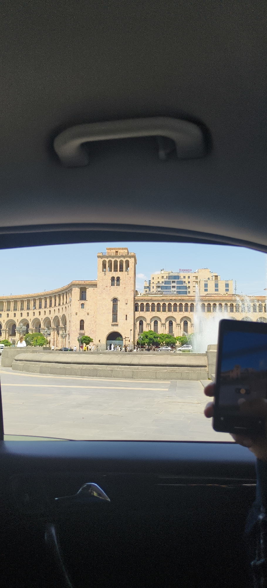This is a photograph taken from inside a vehicle, looking out through the windshield or side window. In the foreground, a dark car interior is visible, including a handle on the ceiling. The main subject of the photograph is a building complex in Yerevan, Armenia, likely Republic Square.
The building is constructed of light-colored stone and features a prominent tower with arched windows. A long, curved colonnade stretches across the facade of the building. In front of the building, there is a public square with a fountain and several people walking. The sky is clear and blue.
To the right of the frame, a person's hand is holding a smartphone, which is capturing the scene. The reflection on the smartphone screen shows a partial view of the building. In the background, a more modern building with glass windows is visible. The overall scene depicts a sunny day in a prominent urban area of Yerevan.
