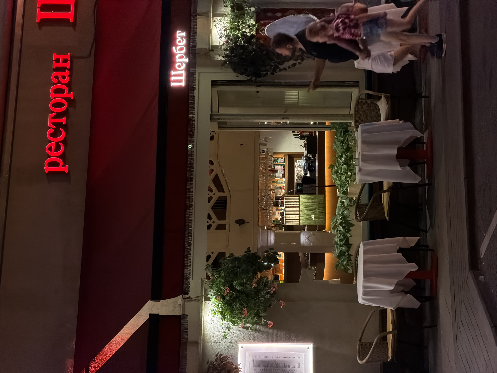The image shows the exterior of a restaurant at night, likely in Moscow, Russia. Red neon letters on the building spell out "ресторан" vertically on the left and "Шербет" vertically on the right. The restaurant has a red awning and a large glass entrance. Outside the restaurant, tables are set with white tablecloths, and chairs are arranged around them. People are visible in the background, some appearing to be staff and others patrons. Green plants are placed around the entrance and on the tables, adding a touch of greenery to the scene. The lighting suggests it is nighttime, and the overall atmosphere appears to be lively and inviting. One table has a small pink flower display.