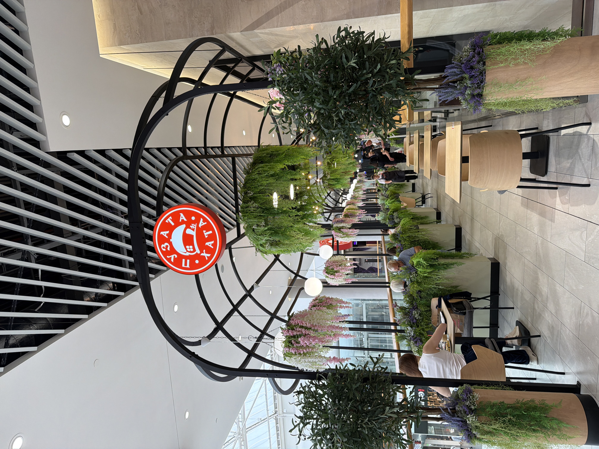 The image shows the interior of a brightly lit cafe, possibly located in the city of Kyiv, Ukraine, given the Cyrillic lettering on the sign. The cafe features a modern design with a black metal arched structure overhead, from which hang artificial plants, including ferns and clusters of pink and white flowers. A prominent circular red sign with white Cyrillic text and a depiction of a house with a hat is suspended from the structure.
Several tables and chairs are arranged in the seating area, with some occupied by patrons. In the foreground, a woman wearing a white tank top and dark pants is seated at a table, looking down at a notebook or tablet. To her right, a man with gray hair is also seated, facing away from the camera. In the background, several servers wearing black aprons are visible, attending to customers or moving through the cafe.
The flooring is tiled, and the walls are clad in a neutral-toned material. Large potted plants with lush green foliage and hanging vines are placed strategically throughout the space, adding to the restaurant's ambiance. The overall environment suggests a contemporary and welcoming cafe setting.