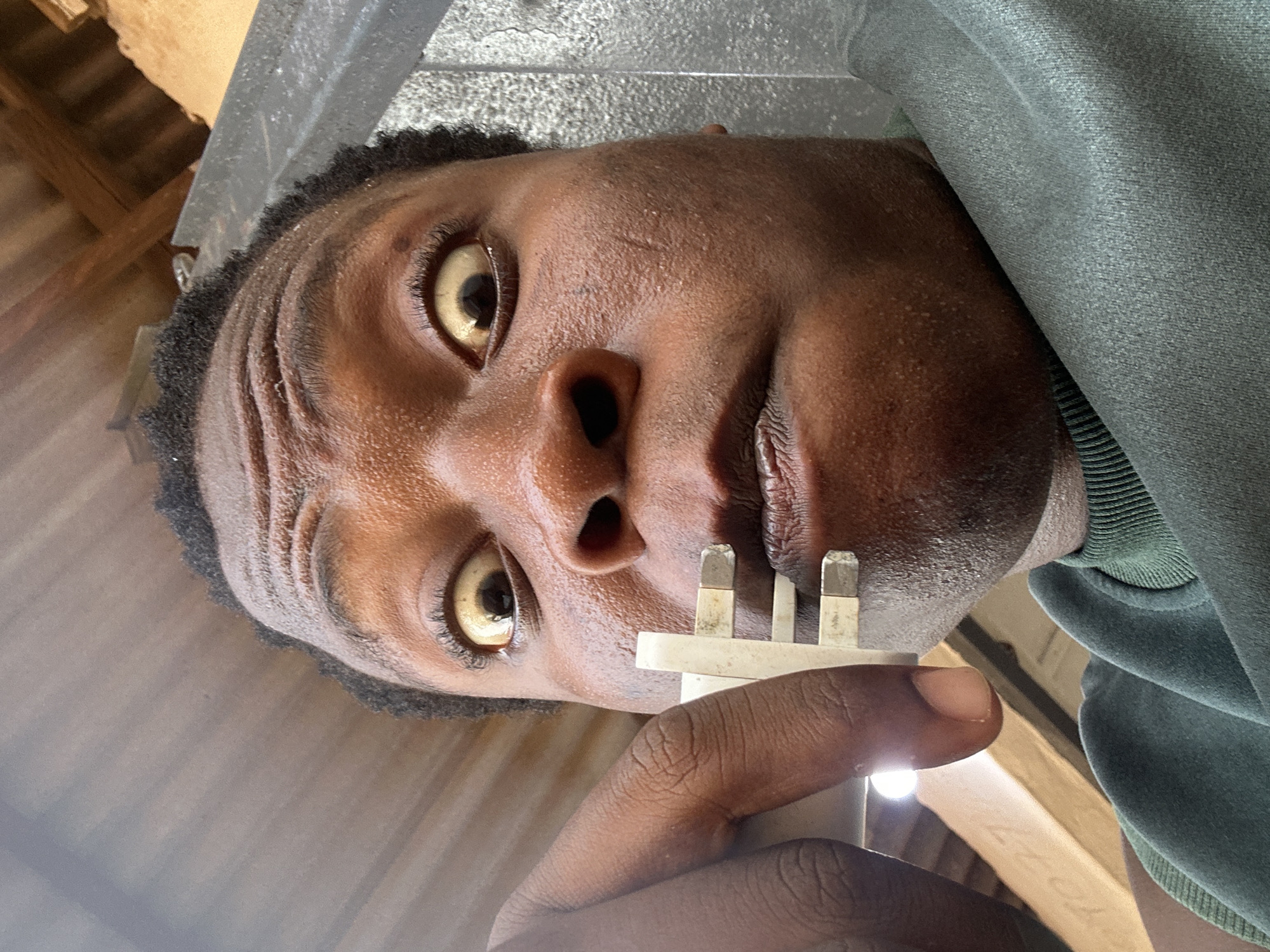 A close-up, low-angle shot shows a person of African descent holding a three-pronged power plug close to their face. The person's eyes are wide, and the plug is positioned near their mouth. The background includes what appears to be interior elements of a building, possibly a ceiling or roof structure. The image's location is provided as Unknown, Unknown.
