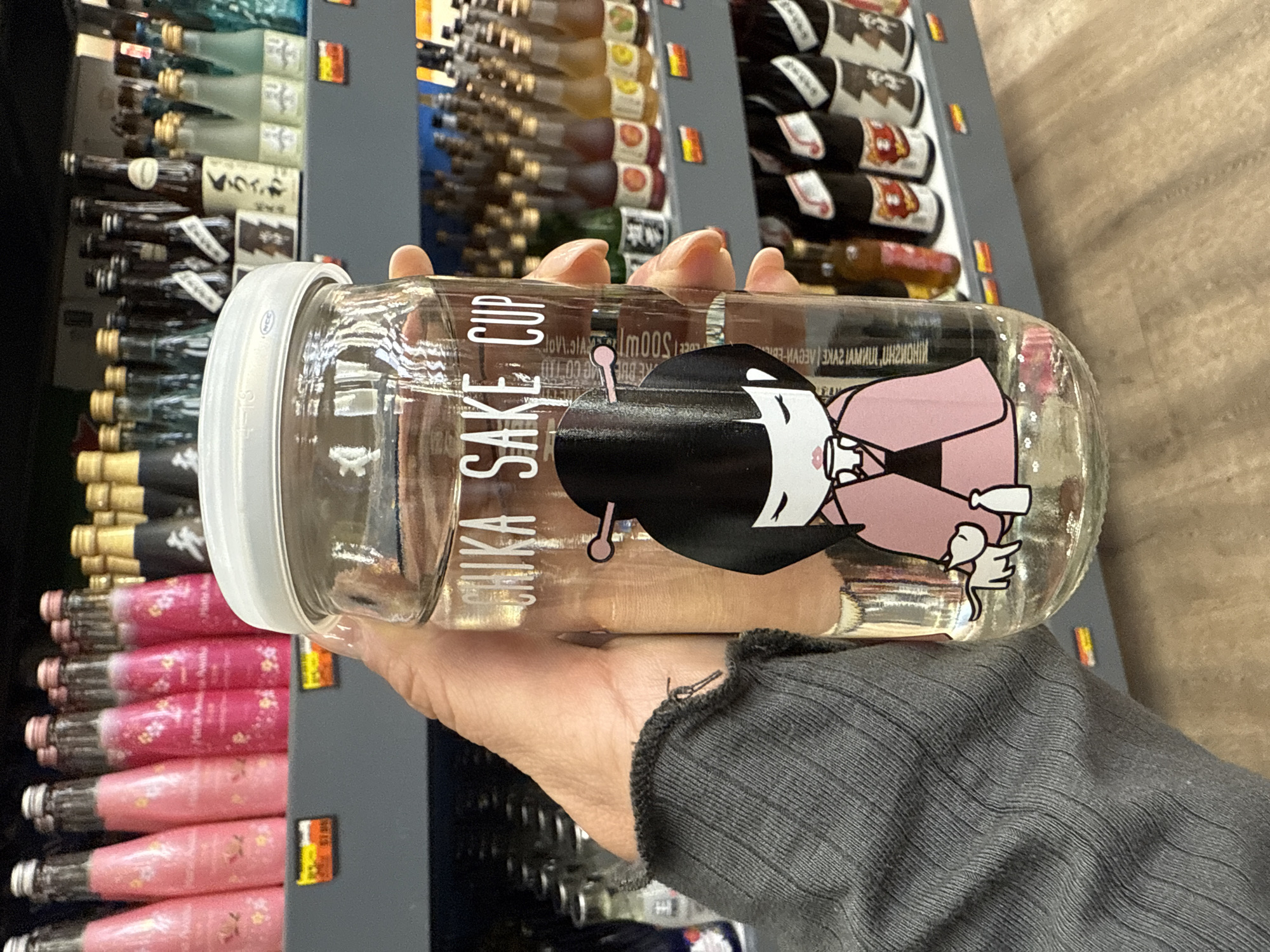 A hand holds a clear glass bottle with a white lid, labeled "CHIKA SAKE CUP," decorated with a cartoon geisha. The background reveals a store aisle in Davie, Florida, with shelves stocked with various bottles of alcohol.