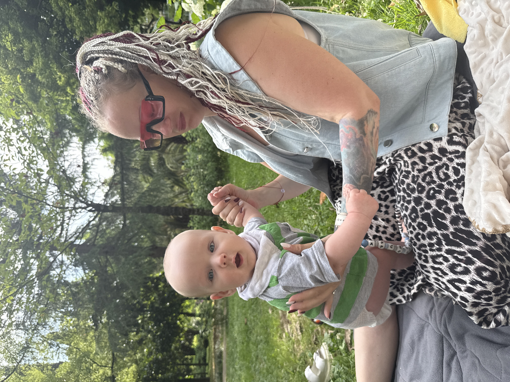 A woman with long, braided hair, featuring streaks of white and burgundy, is holding a baby. She is wearing a light blue denim vest over a leopard-print top. The baby, dressed in a gray and green striped onesie, is looking up at the camera with a surprised expression. The woman's arms are tattooed. They are outdoors in a grassy area with trees in the background, suggesting a park or backyard setting. The lighting indicates it is daytime, likely sunny. A white blanket and a yellow item are visible to the side, possibly picnic items.