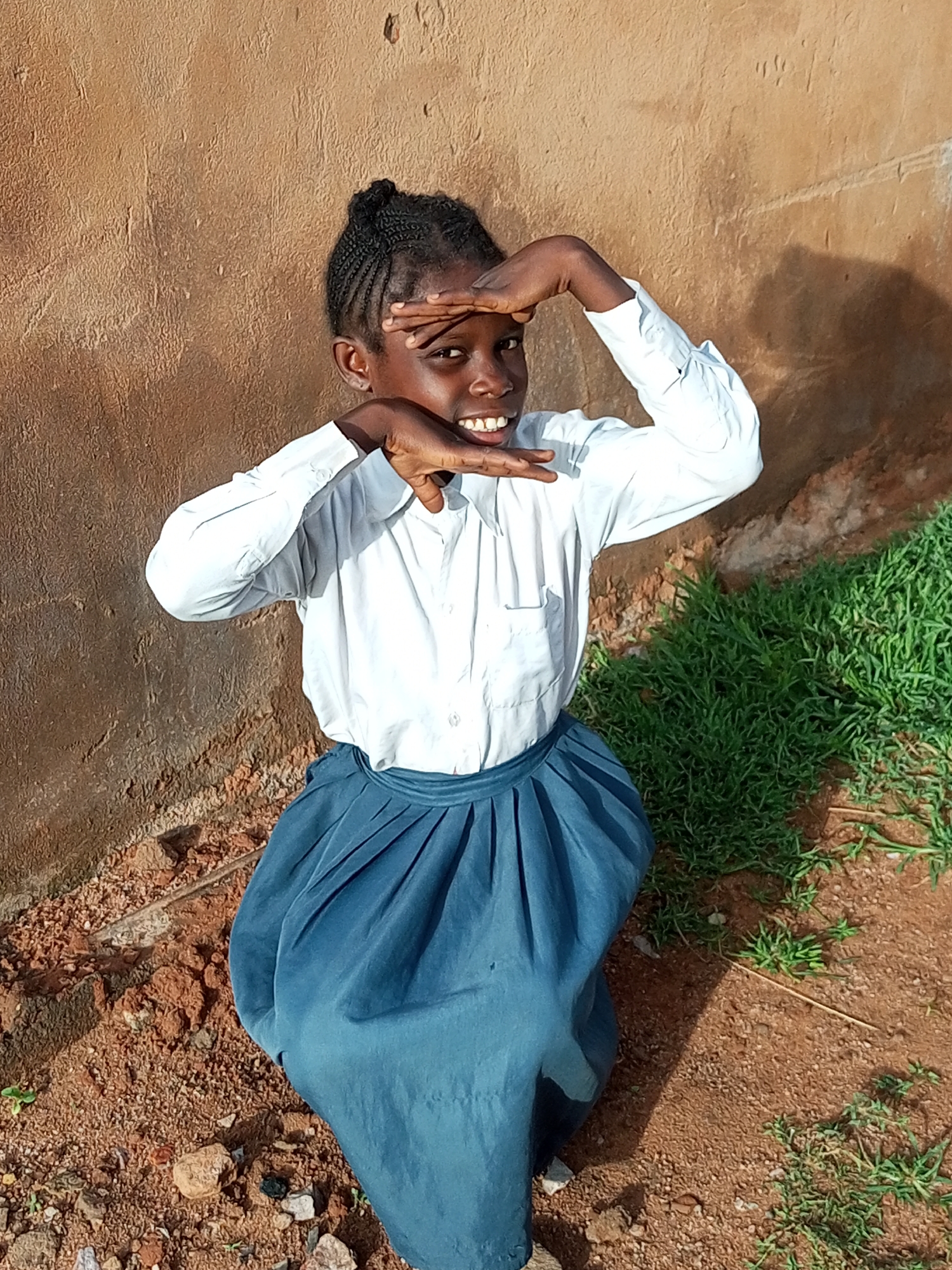 A young Black girl with braided hair pulled into a bun kneels in front of a textured brown wall in Tempure, Nigeria. She is wearing a white collared shirt and a blue pleated skirt. Her hands are held up to her eyes, as if shielding them from the sun, and she is smiling broadly. To her right, a patch of green grass grows from reddish-brown soil. The ground around her is covered in small stones and dirt.
