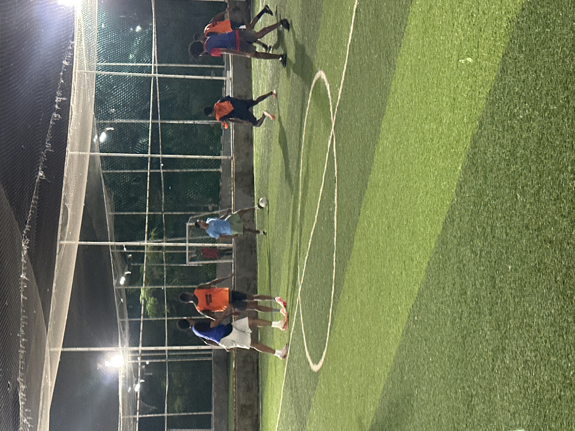 A group of young men are playing soccer on an artificial turf field under artificial lighting, likely in Abuja, Nigeria. The field is enclosed by netting and fencing, typical of urban sports facilities. There are at least six players visible, engaged in a game. One player in a blue shirt and green shorts is actively dribbling a soccer ball. Other players are in motion, wearing orange and blue bibs over their clothing, suggesting a casual game or practice. The lighting creates distinct shadows on the field. The background shows a dark, wooded area beyond the fence, indicating the facility is in a somewhat developed but still green area.