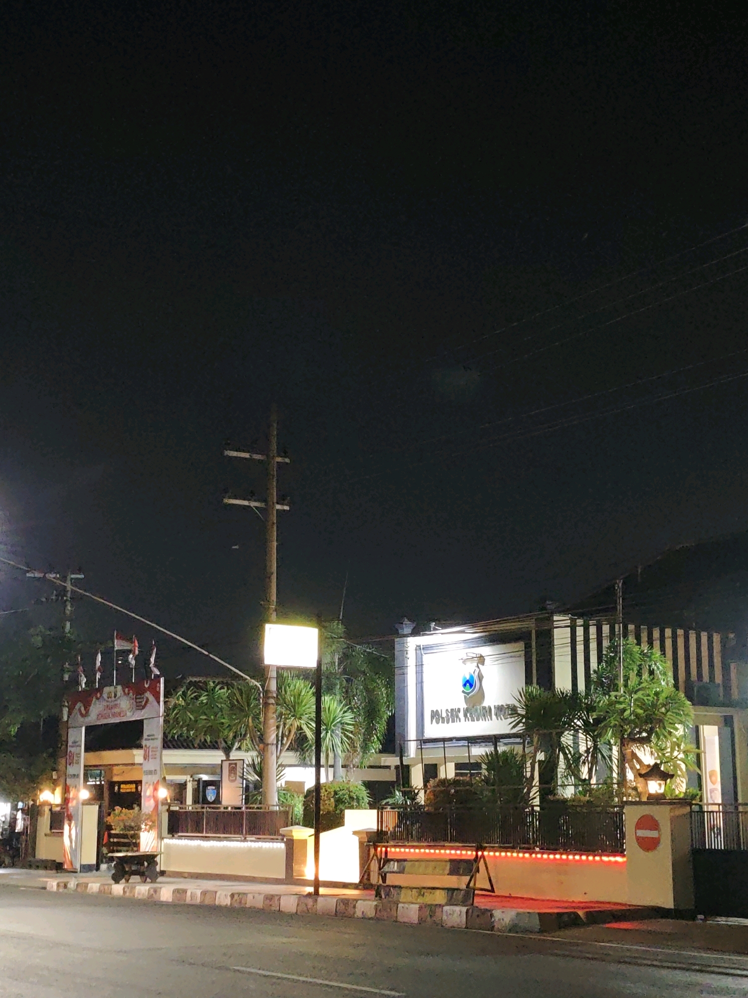 This is a nighttime photograph of the POLSEK Kediri Kota police station in Kediri City, Indonesia. The building is illuminated, with a large, brightly lit sign above the entrance that reads "POLSEK KEDIRI KOTA". The architecture features a modern facade with vertical lines and a prominent logo. In front of the building, there is a fence and some landscaping with trees and plants. Red lights line the bottom of the fence, creating a warm glow.
To the left of the police station, there is an arched gateway decorated with Indonesian flags and banners. A street with a sidewalk and road markings is visible in the foreground. A utility pole with wires stands to the left of the building. The sky is dark, indicating it is nighttime. The overall scene depicts a well-lit and presumably active government building at night, with no people visible in the frame. The atmosphere is calm and orderly.
