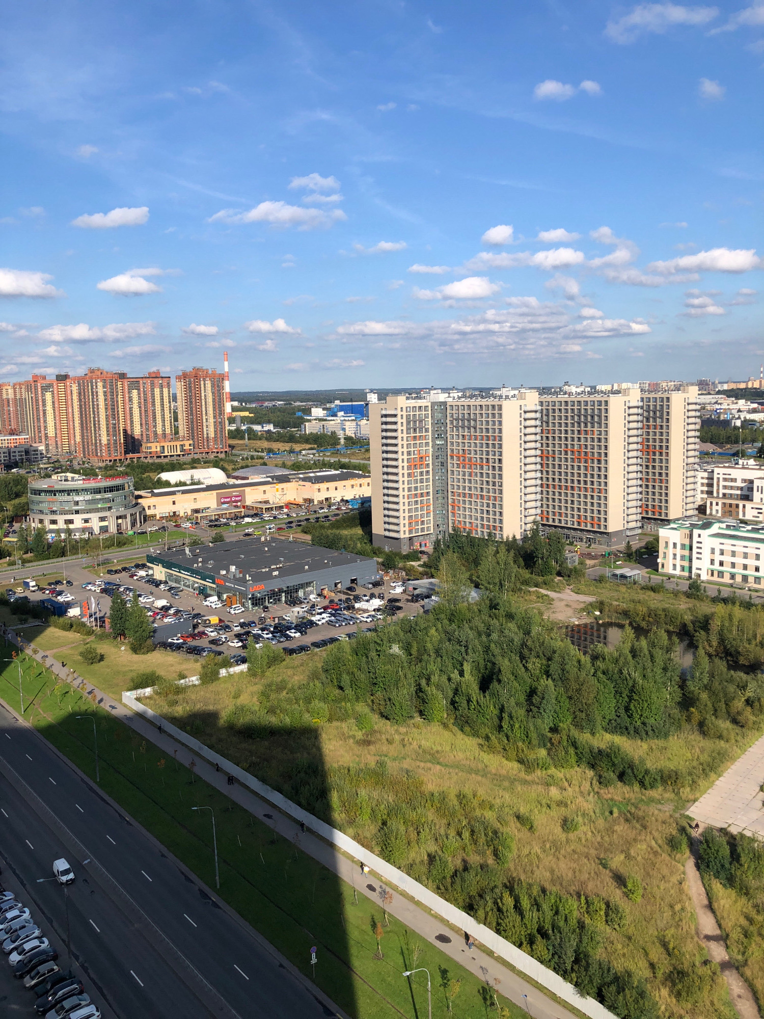 An aerial, medium-distance shot shows a modern urban landscape under a bright blue sky with scattered white clouds. In the background, several large, tan residential buildings with orange accents line the horizon. To the left, two colossal, rectangular residential buildings with light-colored facades and prominent orange vertical stripes dominate the skyline. In the middle ground, a large, modern shopping complex is visible, with a circular building to its left. In front of the shopping complex and to its right, a car dealership with a gray roof displays numerous vehicles, with a prominent "LADA" sign visible. A road runs from the bottom left corner of the frame, with parked cars along its left edge. A white van is driving on the road. The right side of the image features a dense area of green trees bordering a small pond or marsh. The overall scene depicts a developed residential and commercial area, likely in the city of Lavriki, Russia, given the architectural style and context.