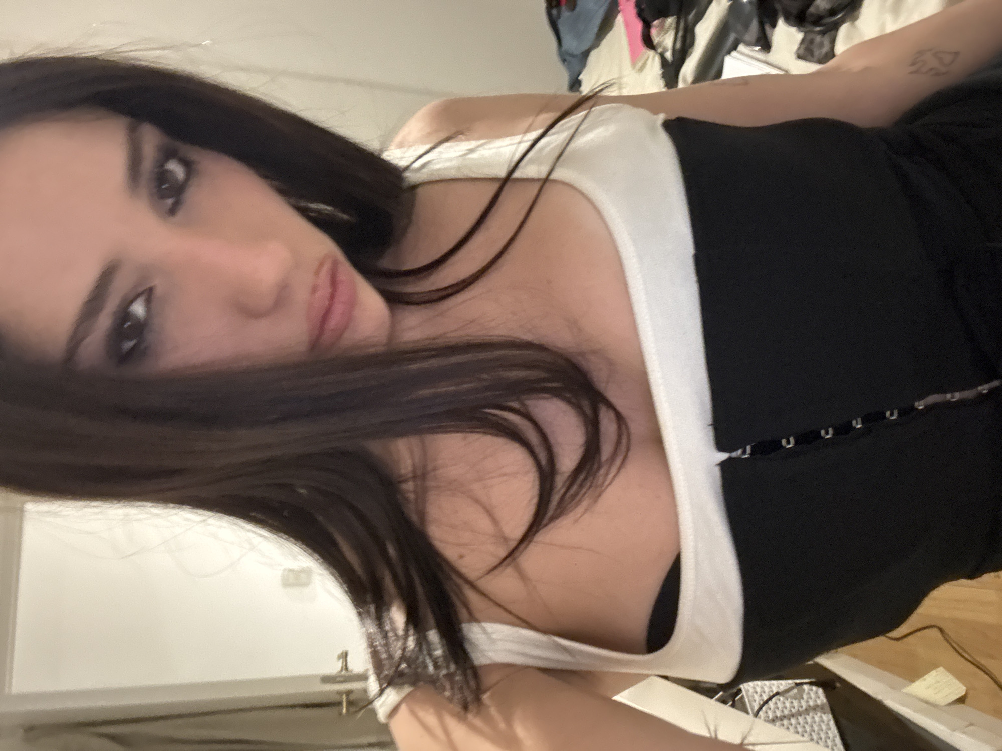 A medium-shot, indoor photograph depicts a woman with dark hair. She is wearing a black and white top and appears to be posing for the camera. The background is cluttered with clothing and other objects. The location cannot be determined from the image.