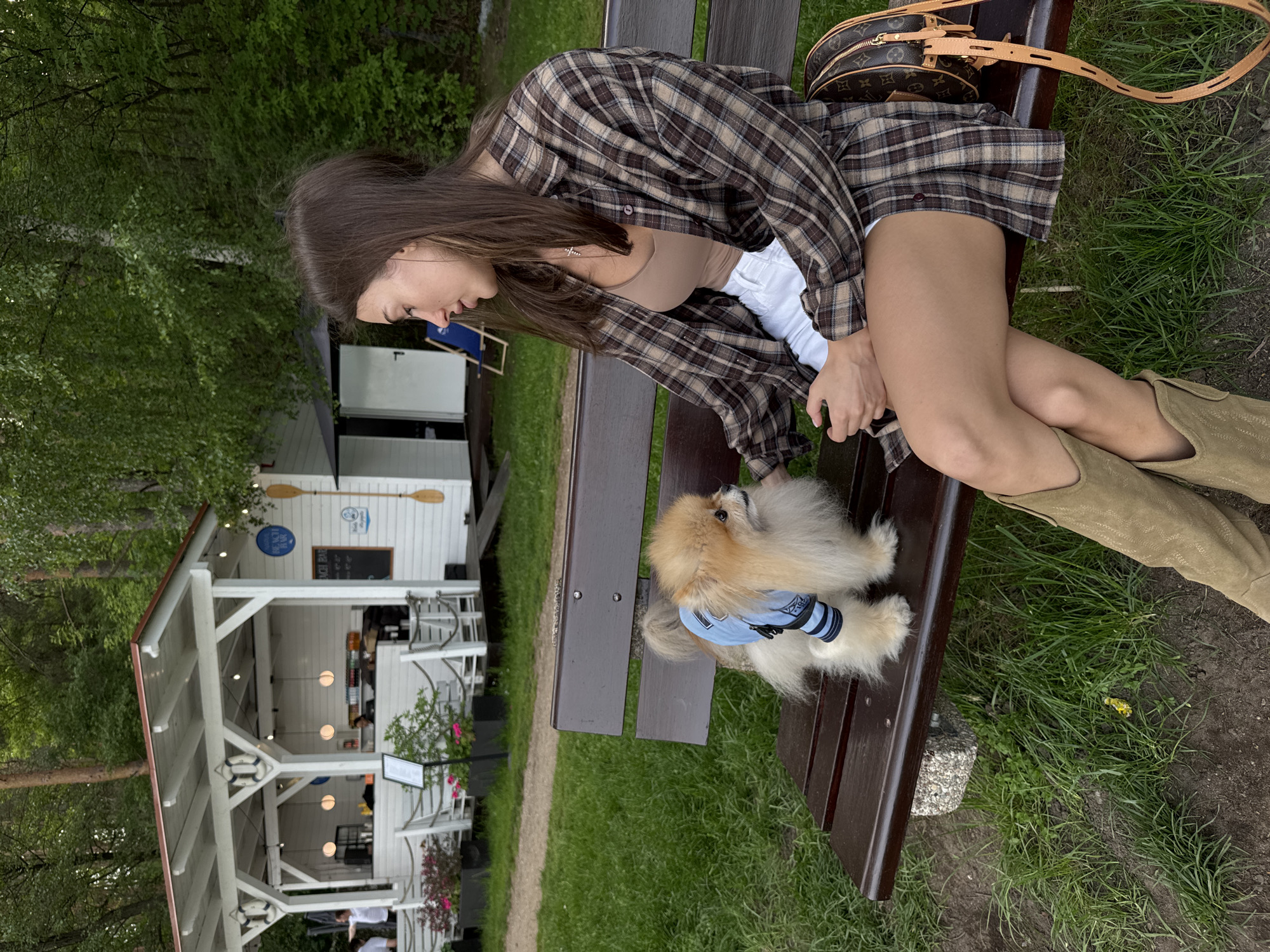 A side-view shot shows a young woman and a Pomeranian dog sitting on a bench in front of an outdoor restaurant. The woman, positioned on the right side of the frame, has long brown hair, a brown tank top, white shorts, and a brown and beige plaid shirt. She is wearing light tan-colored cowboy boots. A small brown handbag sits on the bench next to her. The dog, wearing a light blue shirt with dark blue stripes, is seated between the woman's leg and the back of the bench. The bench is made of dark brown wood.
In the background, a white building with an open structure and a dark-colored roof is visible. Tables and chairs can be seen inside the building, suggesting it is a restaurant or outdoor dining area. Trees and green foliage surround the building, adding to the outdoor setting. Patrons can be seen at the tables inside the building. The location is in a grassy area.