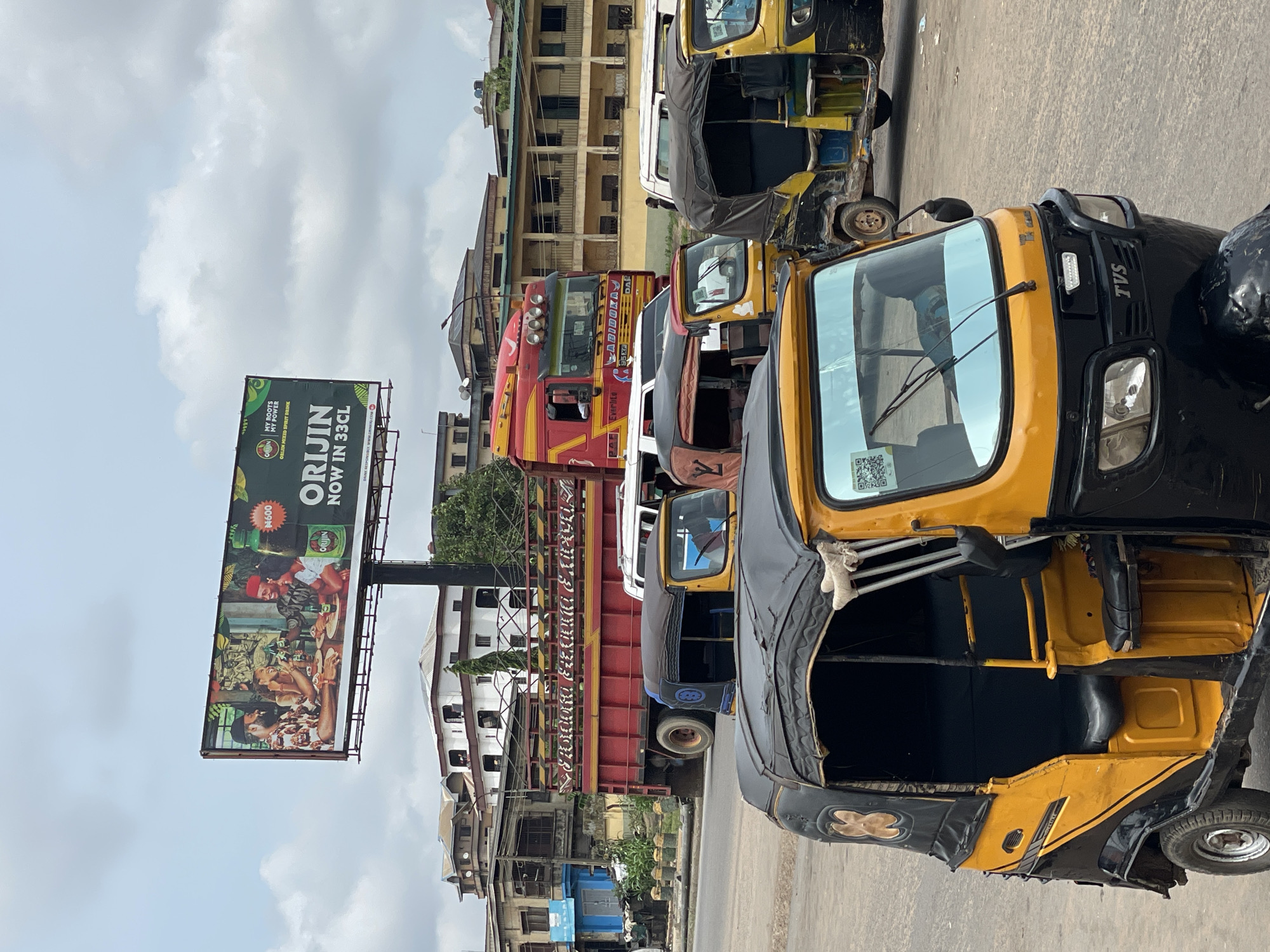 The image depicts a street scene in Onitsha, Nigeria, characterized by a row of yellow and black auto-rickshaws (Tuk-Tuks) parked along the roadside. A red and yellow truck with the word "LABIDOKAY" painted in white stands behind the auto-rickshaws. A large billboard advertising "ORIJIN" drink is visible in the background. The buildings along the street are multi-storied and appear to be a mix of residential and commercial properties. The sky is overcast with cloudy conditions. The foreground is dominated by the auto-rickshaws, while the background showcases the urban landscape of Onitsha. The overall scene conveys a typical day on a busy street in this Nigerian city.