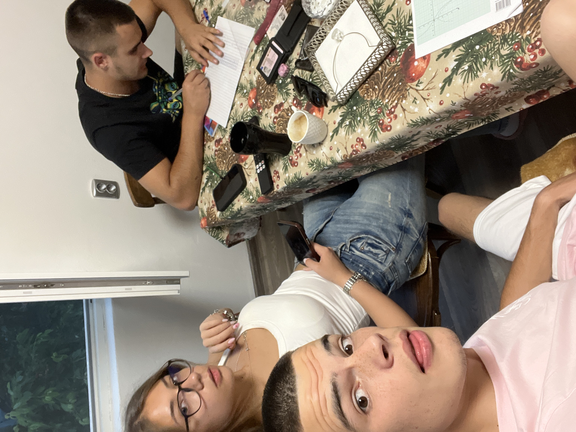 This image captures an indoor scene in Karavelovo, Bulgaria, featuring three young adults. In the immediate foreground, a male with short dark hair, freckled skin, and wearing a pink t-shirt, looks toward the camera with his tongue slightly extended. His right eye is visible. Behind him and to his left, a female with long brown hair, dark-framed glasses, a white tank top, and blue jeans, sits while looking directly at the camera. She holds a thin silver necklace with her right hand near her chest and a dark smartphone in her left hand, which rests on her lap. A silver watch is on her left wrist. To her right, a second male with short dark hair, wearing a black t-shirt with a colorful graphic and a silver chain, is seated at a table, bent forward, and actively writing on a piece of lined paper with a pen.
The table is covered with a festive tablecloth patterned with pinecones, red berries, and green foliage. Various objects are distributed across its surface: a dark smartphone, a black remote control, a black tumbler, a white coffee mug with residual liquid, black sunglasses, and a decorative silver tray containing a napkin holder. Near the writing male, an open black wallet displaying documents, a small pink circular object, and a red case are visible. Another sheet of paper with a green border and a plotted graph is partially visible on the right edge of the table.
The background consists of a plain white wall. A window on the left side reveals green foliage outside. A vertical white window frame or blind mechanism is present beside the window. A grey electrical outlet with two circular sockets is mounted on the wall to the right. Dark wooden chairs are partially visible behind the two individuals seated at the table. The floor is dark, with a light-colored, possibly woven, object under the foreground male's foot.