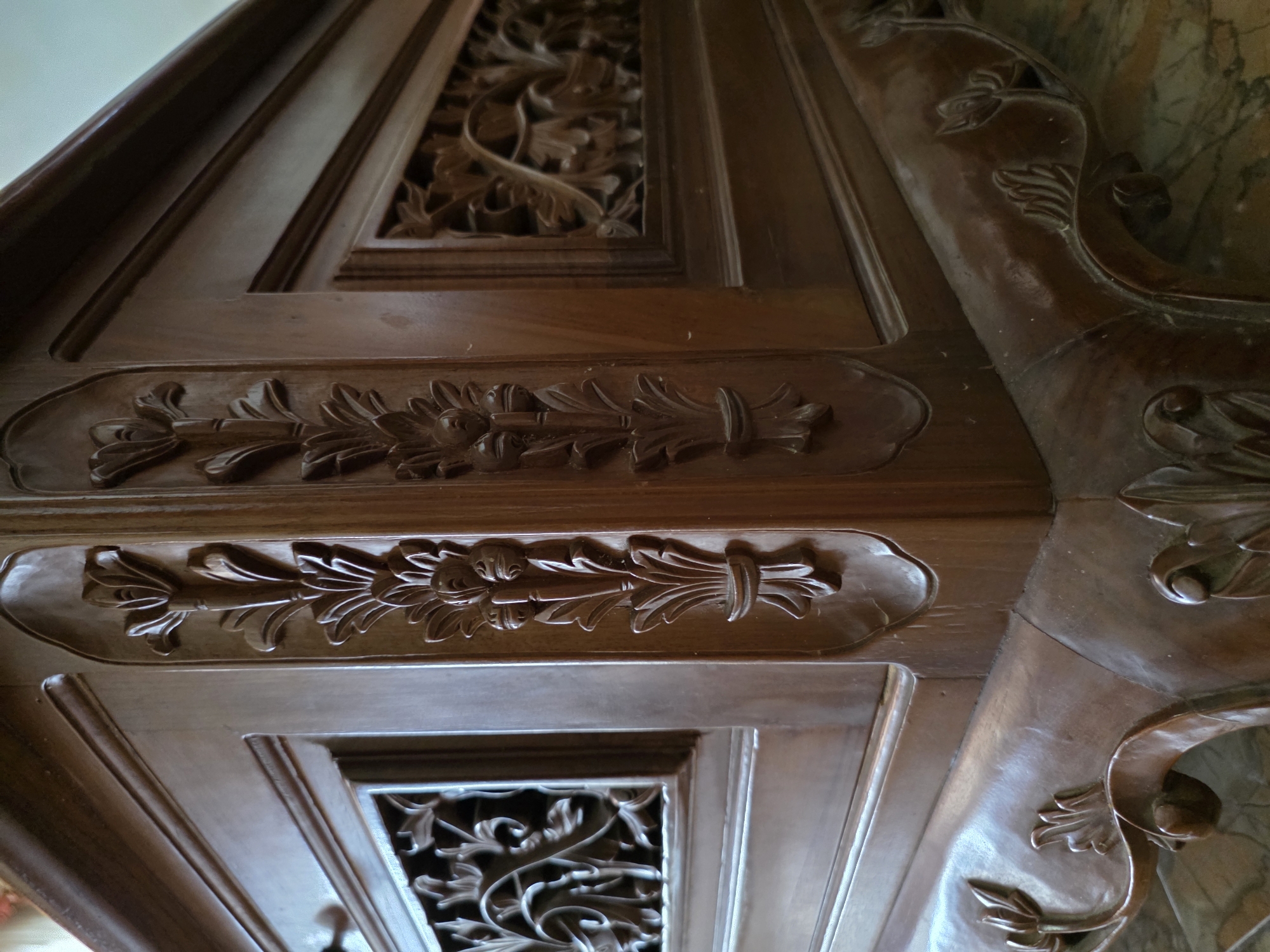 The image shows a detailed close-up of carved wooden furniture, likely a table or cabinet, with intricate floral motifs. The wood is a dark, rich brown and appears to be polished. The carvings are deep and elaborate, featuring stylized flowers and leaves. The furniture has a prominent corner with detailed decorative elements. The background is out of focus, but suggests an indoor setting with a marble or stone floor. There are no people visible in the image. The scene is static and focuses solely on the craftsmanship of the wooden object. There is no information or context provided within the image to suggest a specific location in Bireuenmeunasahblang, Indonesia.