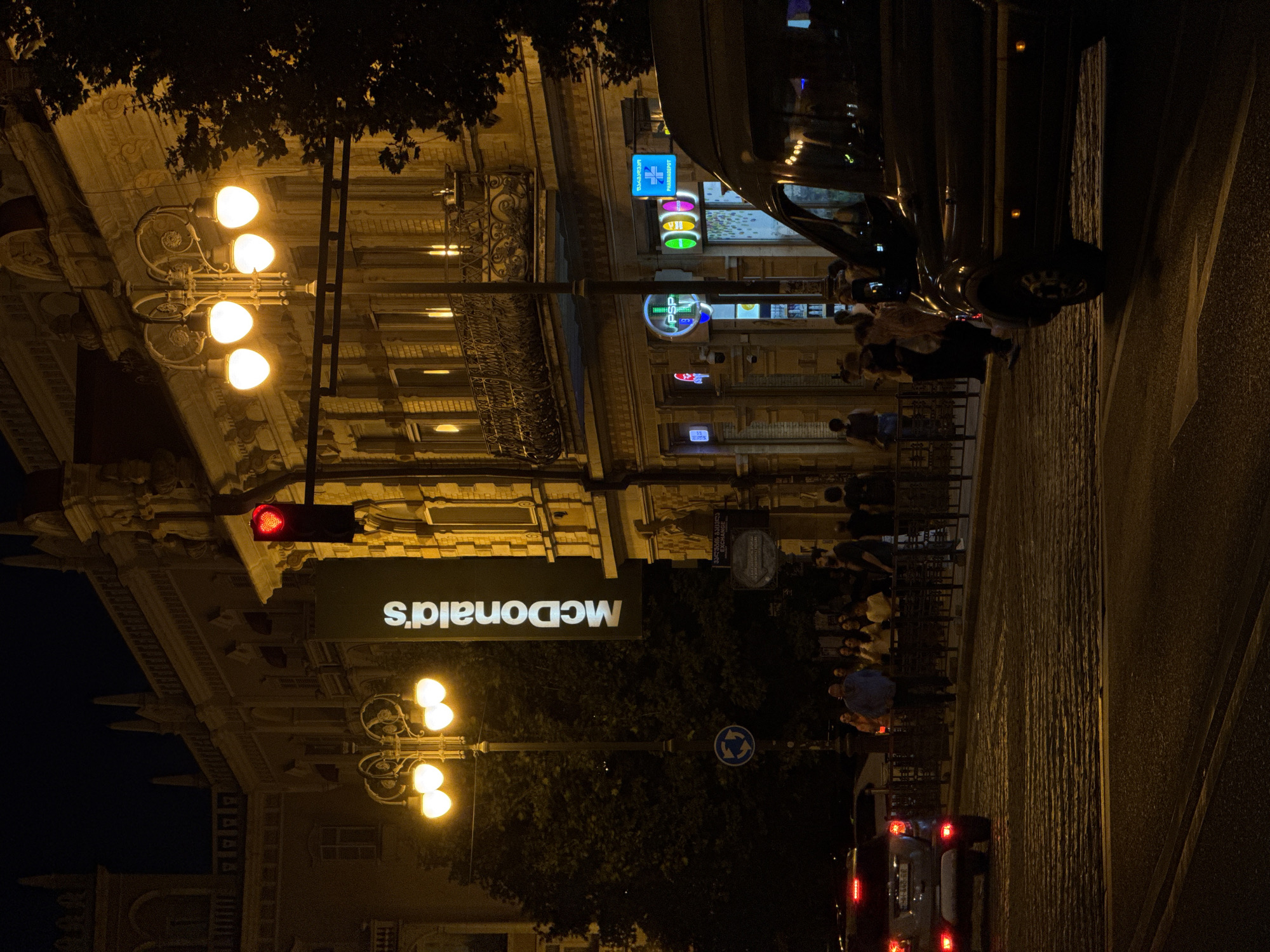 At night, on a cobblestone street in Tbilisi, Georgia, a red heart-shaped traffic light is illuminated. A large McDonald's banner hangs on the side of a building. In the foreground, a dark-colored van is parked at an intersection. On the sidewalk, people are walking and standing, some are gathered near a fence. A silver car is driving away from the viewer. Streetlights and illuminated shop signs cast a warm glow on the scene.