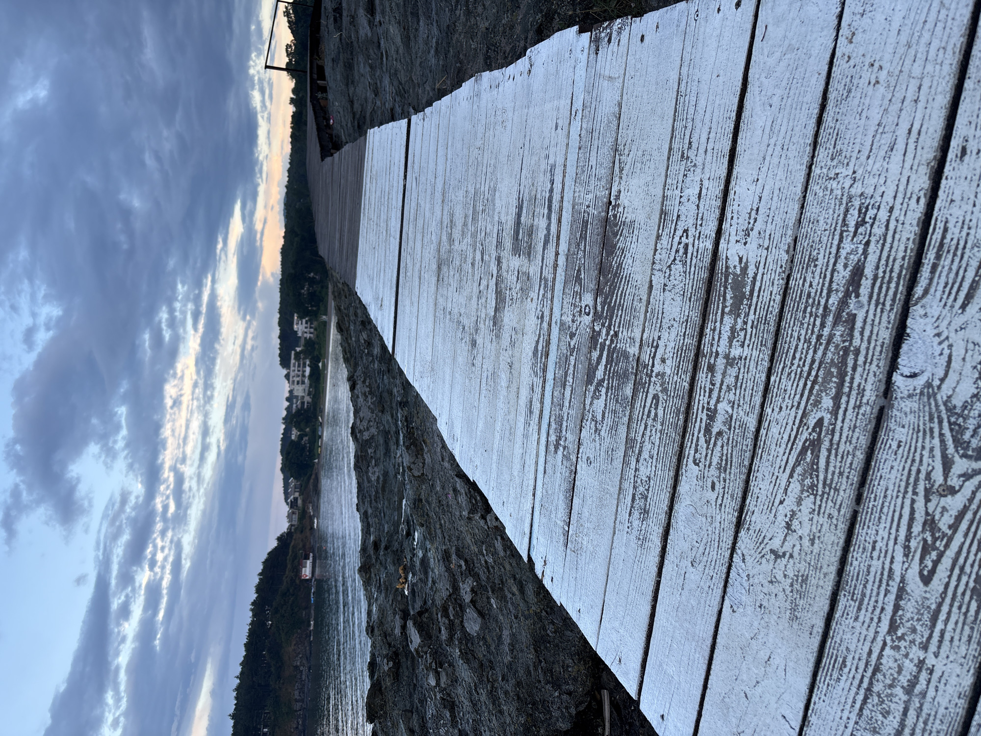 A weathered, whitewashed wooden boardwalk leads toward a body of water. The boardwalk is in the foreground and dominates the lower half of the image, showing signs of wear with chipped paint revealing the dark wood beneath. To the left of the boardwalk, dark, rocky terrain slopes down towards the water. The water itself is calm, reflecting the muted colors of the sky. In the distance, across the water, is a coastline with a line of trees and several buildings, including what appears to be a cluster of houses or small hotels. The sky above is filled with dramatic, dark clouds tinged with the soft light of dawn or dusk. The overall scene suggests a coastal location, possibly a resort area, in a somewhat overcast and tranquil atmosphere. The scene is located on the coast of Tsarevo, Bulgaria.