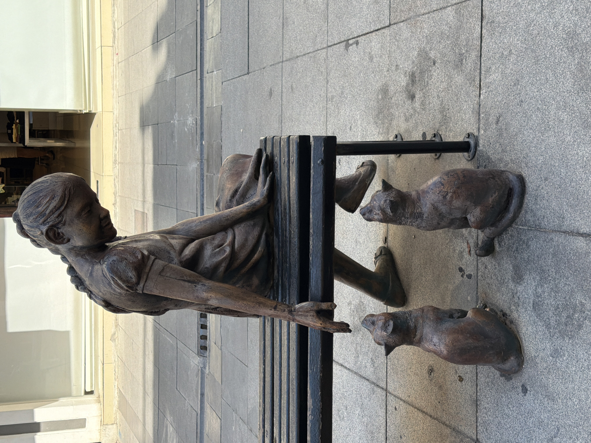 A bronze statue depicts a young girl with her hair in a braid, wearing a dress, sitting on a dark wooden bench. Her head is bowed, and her right hand is extended towards two small bronze cat statues positioned on the pavement below. The girl's left leg is bent, with her foot resting on the ground, while her right foot is also on the ground. The scene is set on a paved sidewalk in front of a building with large windows. The lighting suggests it is daytime, with shadows cast on the ground. This sculpture is located in Marmaris, Turkey.