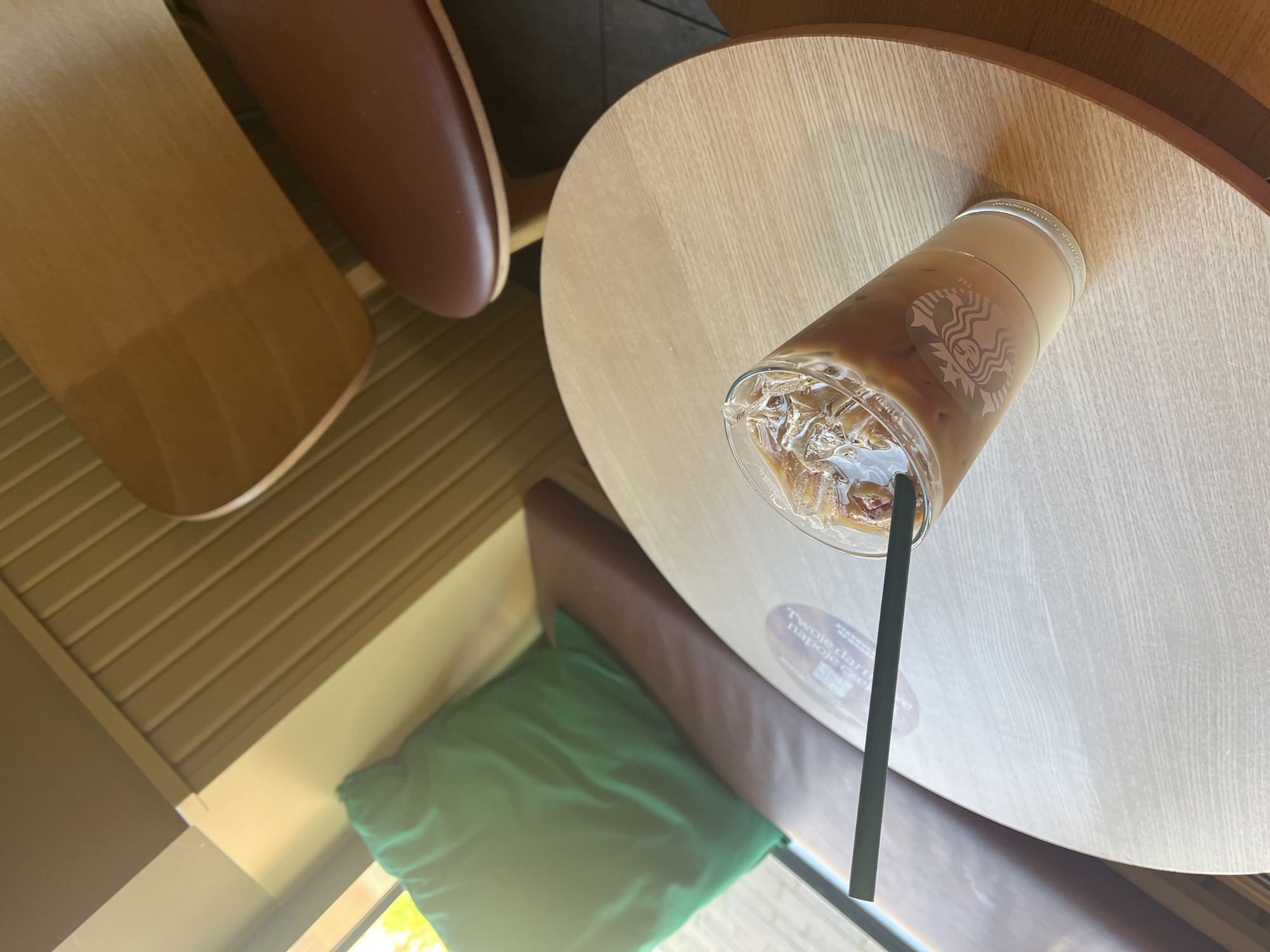 An iced coffee with a black straw sits on a circular wooden table in a cafe. The coffee cup has the Starbucks logo visible. A cushioned bench with a green pillow is to the left, and two wooden chairs with brown seats are in the background. The walls are paneled with vertical wood slats. Sunlight streams in from a window on the left side of the image.