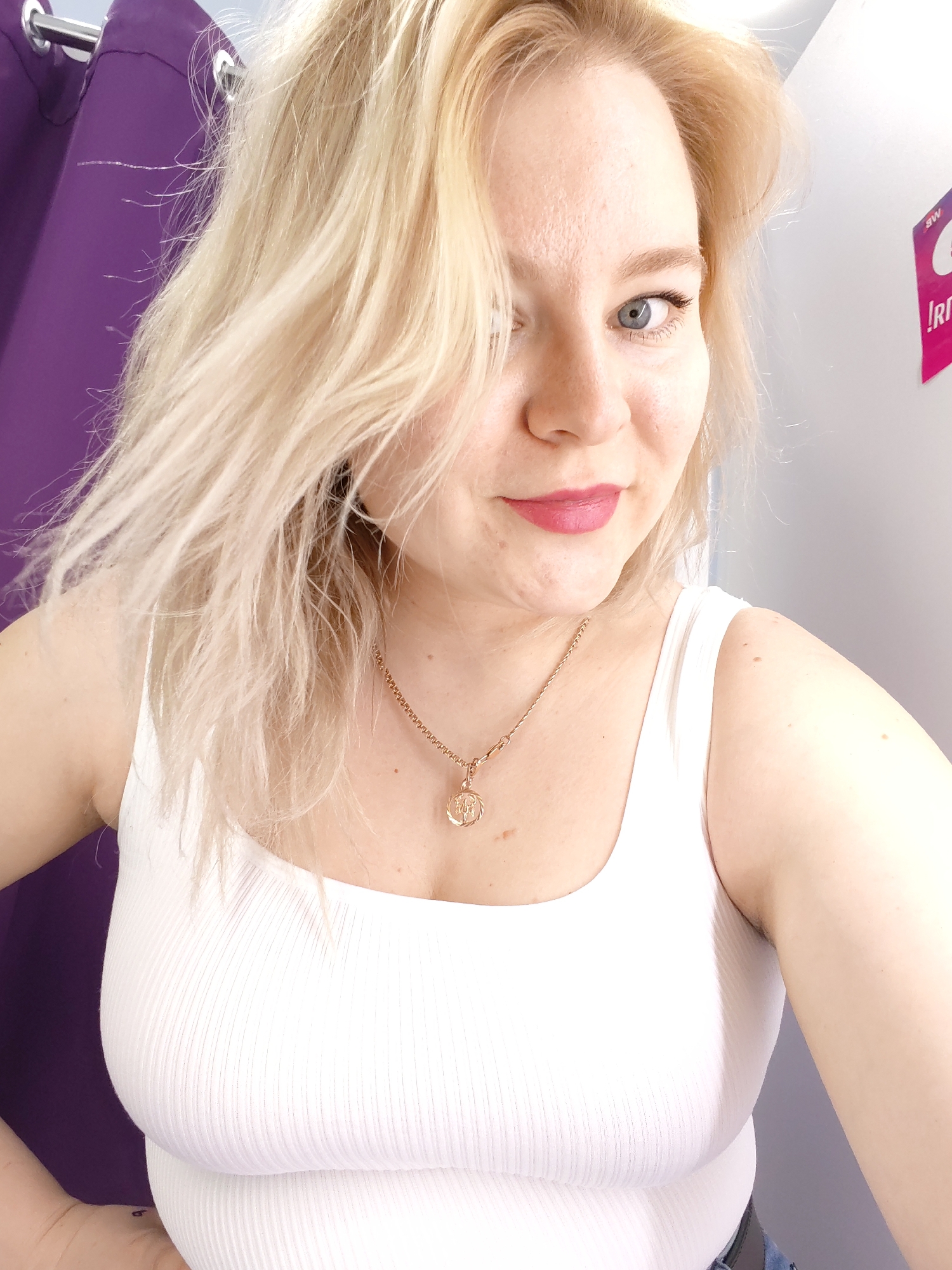 A fair-skinned woman with blonde hair and blue eyes is posing for a selfie in what appears to be a fitting room. She's wearing a white ribbed tank top and a gold necklace with a pendant. The woman has pink lipstick on and is looking directly at the camera with a slight smile.
Behind her, a purple curtain hangs to the left. To her right, a white wall is visible with a pink sign bearing an inscription. The location is in the city of Izhevsk, Russia.