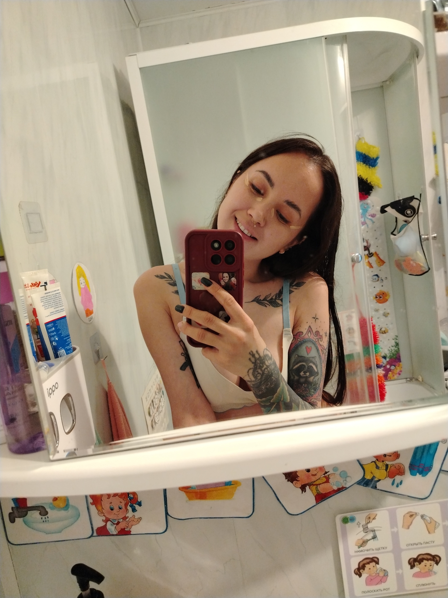 A selfie is captured in a bathroom mirror, reflecting a woman with dark hair. She is holding a burgundy cell phone. She has fair skin, a nose ring, and light brown strips are placed under her eyes. Tattoos are visible on her arms and chest. The bathroom has a white sink and walls. The lower portion of the mirror shows a white shelf decorated with childish cartoons. Toothpaste and an Ippo dispenser sit on the shelf. The bathroom appears to be located in Noviy, Russia, judging from the text on the toothpaste tubes.