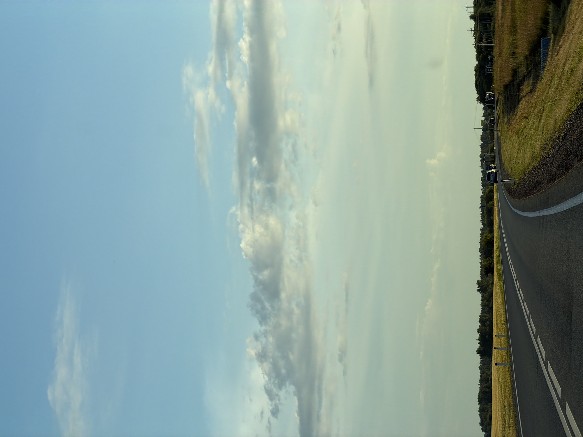 The image depicts a paved road curving through a rural landscape under a sky with scattered clouds. The road has white lane markings. To the left of the road, a field of yellow crops, likely wheat or barley, stretches to a line of trees in the distance. White posts with black tops mark the edge of the road. To the right of the road, tall green grass and some shorter vegetation are visible, with more trees and structures, possibly rural buildings or power lines, further in the distance. A silver car is seen driving on the road in the middle ground, and another vehicle appears to be further ahead. The time of day appears to be late afternoon or early evening, given the soft lighting. The environment is natural and agricultural, suggesting a location outside of a major urban center. The image does not provide any specific identifying features that would pinpoint its location within the city of Aryshkhazda, Russia, or any other specific city.