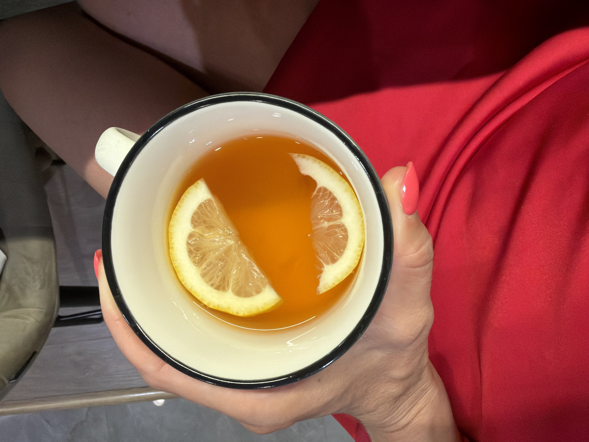 The image presents a top-down view of a person holding a mug, set in an indoor environment in Birobidzhan, Russia. The central subject is a white ceramic mug with a prominent black rim, held firmly by a hand. The hand has a light skin tone, and its nails are painted with a bright pinkish-coral polish. Inside the mug, an amber-colored liquid, strongly suggesting tea or a similar warm beverage, contains two fresh lemon slices. One large lemon slice is positioned on the left side of the mug, while a second, slightly smaller slice floats on the right, contributing to the drink's visual appeal.
To the right of the mug, a significant expanse of a vibrant red fabric is visible. This material appears soft and flowing, characterized by its natural folds and drapes, indicating it is likely part of a dress or another garment worn by the person. On the upper left portion of the frame, a segment of the person's leg or arm, also with a light skin tone, can be seen, partially obscured by shadow. Further to the left and slightly below, a light-colored, possibly beige or off-white, upholstered surface or piece of furniture is discernible. The immediate surface beneath the mug is dark and flat, possibly a table or lap. The scene evokes a sense of comfort and a peaceful, personal moment. There is no visible text in the image.