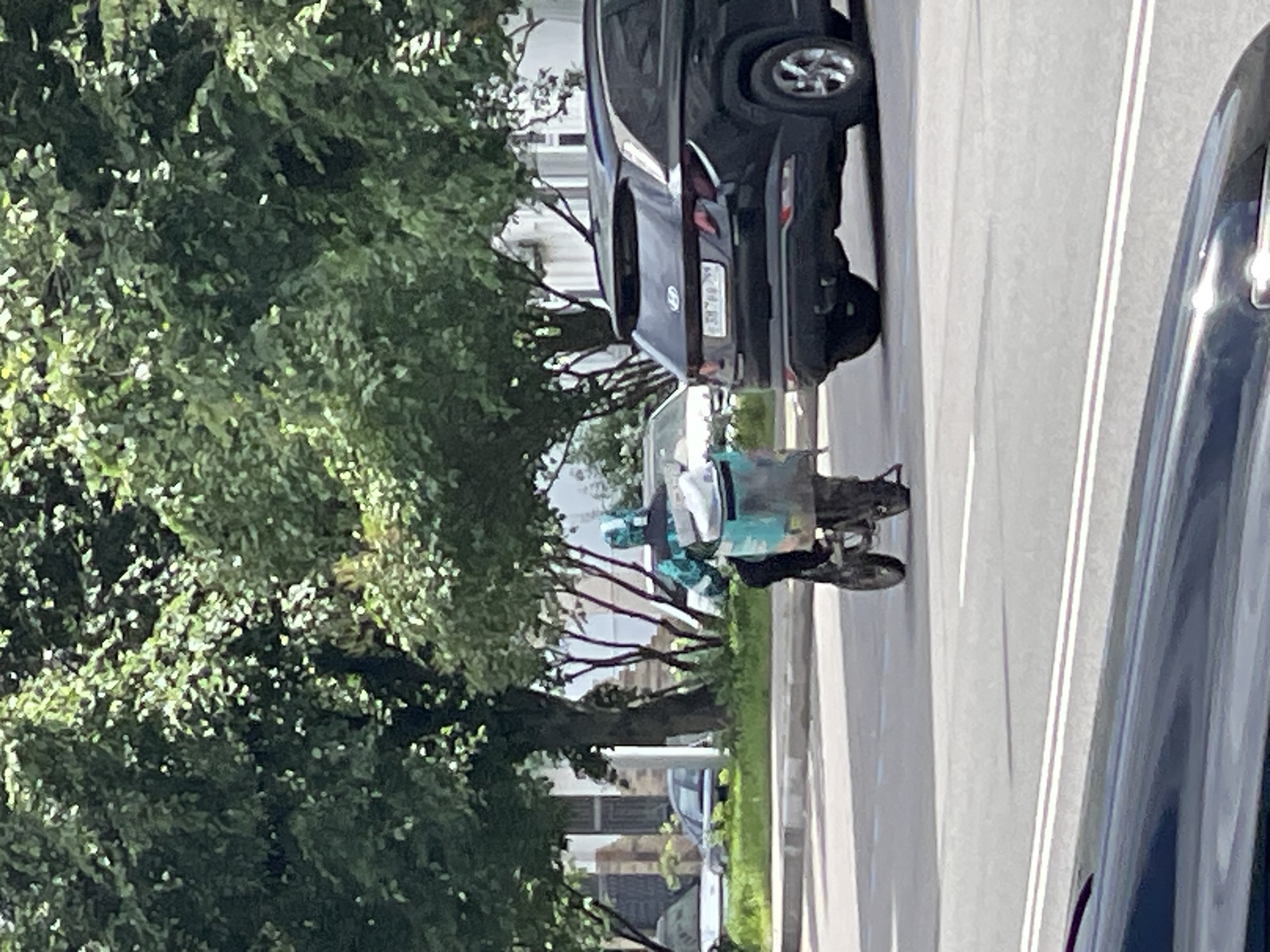 A delivery person on a motorcycle is seen from the rear on a street in Moscow, Russia. The delivery person is wearing a blue and green patterned jacket and a helmet. They are riding a motorcycle with a large, light blue insulated delivery box mounted on the back. The motorcycle is in motion on a paved road, with a black SUV to its right. In the background, there are trees, buildings, and other vehicles. The scene appears to be in an urban environment during daylight hours.