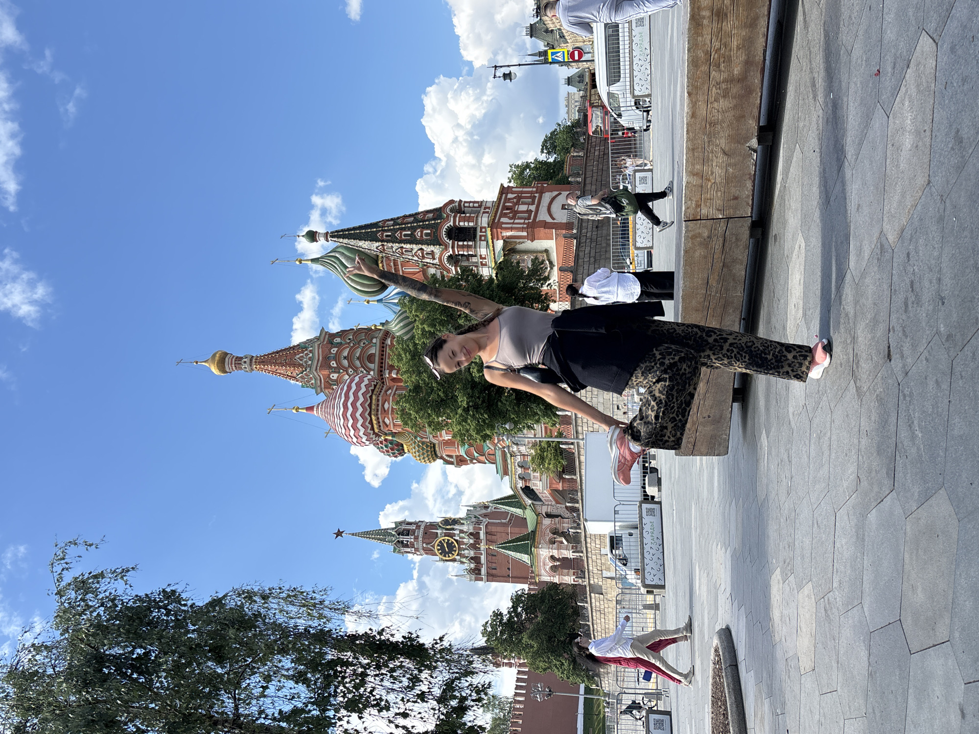 The image shows a woman posing in front of St. Basil's Cathedral in Moscow, Russia. The woman, wearing a tank top, leopard-print pants, and pink sneakers, stands on a stone-paved area, her arm raised towards the cathedral. Behind her is a portion of St. Basil's Cathedral with its distinctive colorful domes. In the background are other buildings, people, a bus, and a blue sky with white clouds. A tree is visible on the left side of the image.