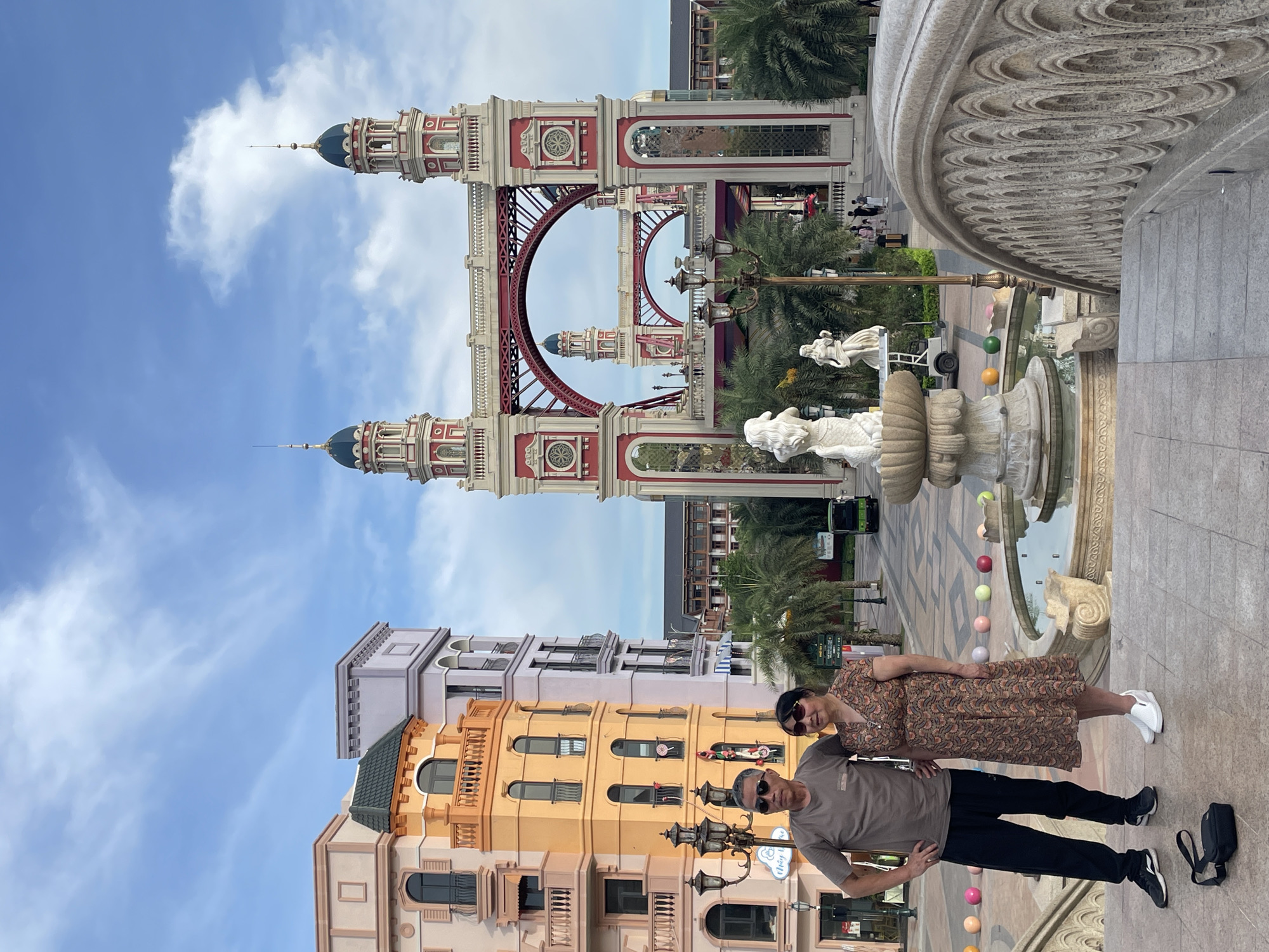 An elderly Asian couple, the woman in a floral dress and the man in a t-shirt and pants, pose for a photo on a stone bridge with intricate carvings. They stand on a patterned tiled plaza in front of a whimsical architectural structure resembling a red and white bridge with two towers. Behind them, colorful buildings with balconies and arched windows line the street, creating a picturesque, European-inspired urban setting. Palm trees are scattered throughout the plaza, and a fountain with sculptures is visible. The sky above is a clear blue with scattered white clouds. In the distance, other pedestrians and a small green vehicle are visible. The overall scene suggests a tourist destination or a themed outdoor space.