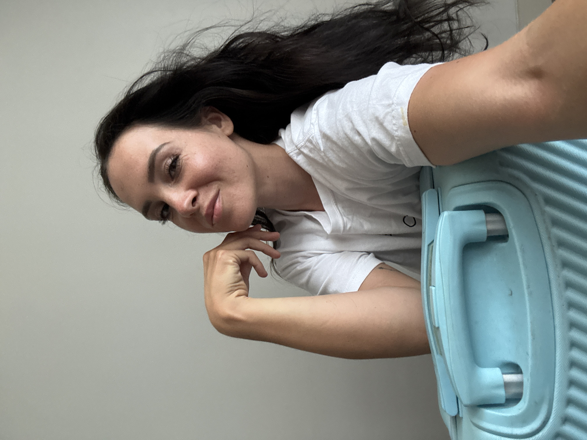 A woman with long, dark hair and a white t-shirt leans on a light blue suitcase. She rests her chin on her hand, looking towards the camera with a slight smile. Her hair is blowing to her right. The background is a plain, light gray wall. The scene appears to be indoors.