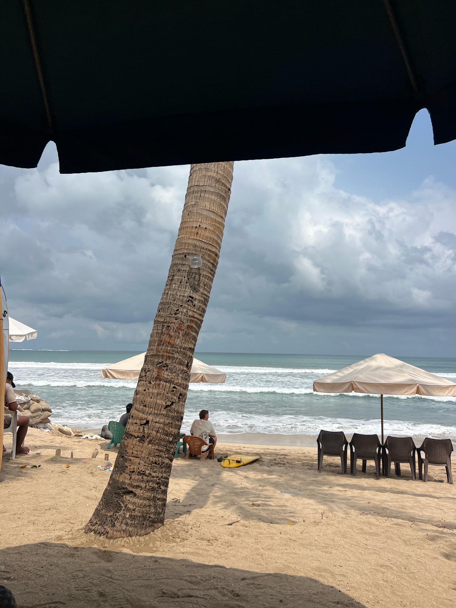 The image depicts a cloudy day at Kuta Beach in Kuta, Indonesia. In the foreground, a large palm tree trunk leans diagonally from the left to the right. A dark umbrella partially obscures the top of the image. The beach sand is light brown, with footprints visible. Several people are seated on chairs, facing the ocean. To the left, a person sits on a chair with sandbags behind them. In the middle ground, two people are seated near the water's edge, one on a green chair and another on a brown chair. Further to the right, under a beige umbrella, four dark plastic chairs are arranged in a row. A yellow surfboard lies on the sand to the left of the chairs. The ocean waves are breaking on the shore, and the sea is a greenish-blue color. The sky is filled with a mixture of white and dark gray clouds.