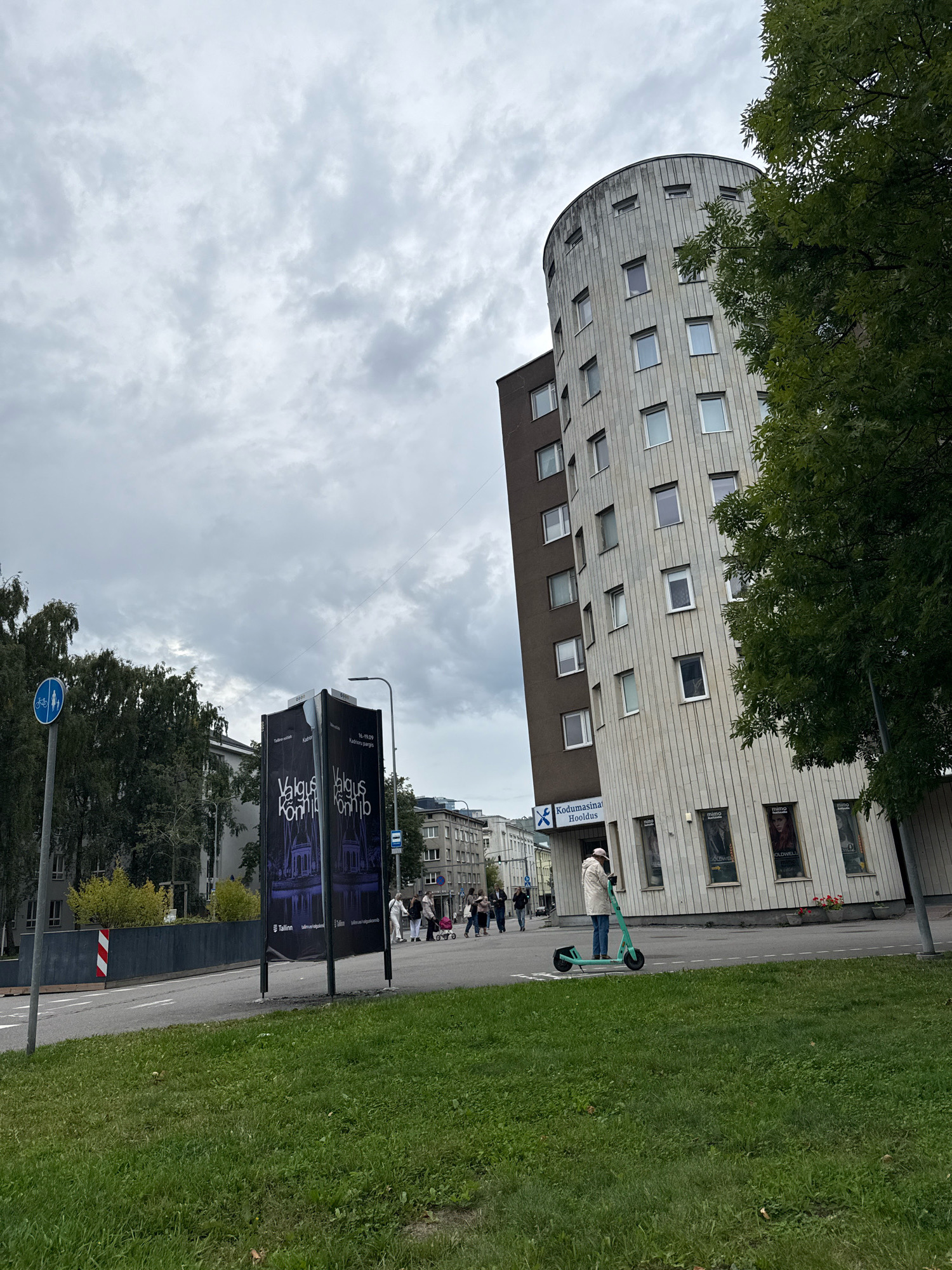 The image depicts an urban scene in Tallinn, Estonia, on a cloudy day. In the foreground, a patch of lush green grass slopes upwards towards a grey asphalt road. Across the road, a modern, cylindrical building with light-colored vertical panels and numerous rectangular windows dominates the right side of the frame. A dark brown section is visible on the left side of this building. Above the entrance of this building, a sign reads "Kodumasina Hooldus" with a blue logo. Several posters are affixed to the lower section of the building's facade, some featuring the word "MIMO" and portraits.
To the left of the building, a dark-colored bus stop shelter is partially visible. In front of the bus stop, a black and white directional sign indicates a bike path and a pedestrian walkway. A tall light pole with a street lamp stands near the bus stop and the bike path sign.
Further left, a large billboard displays images and text, with the words "Valgus Kõnnib" prominently featured. Multiple people are walking in the background near a crosswalk. One person in the middle ground is riding a teal electric scooter. Trees with dark green foliage are present on the left and right sides of the image, with the tree on the right partially obscuring the upper portion of the building. The sky is overcast with layers of grey clouds.