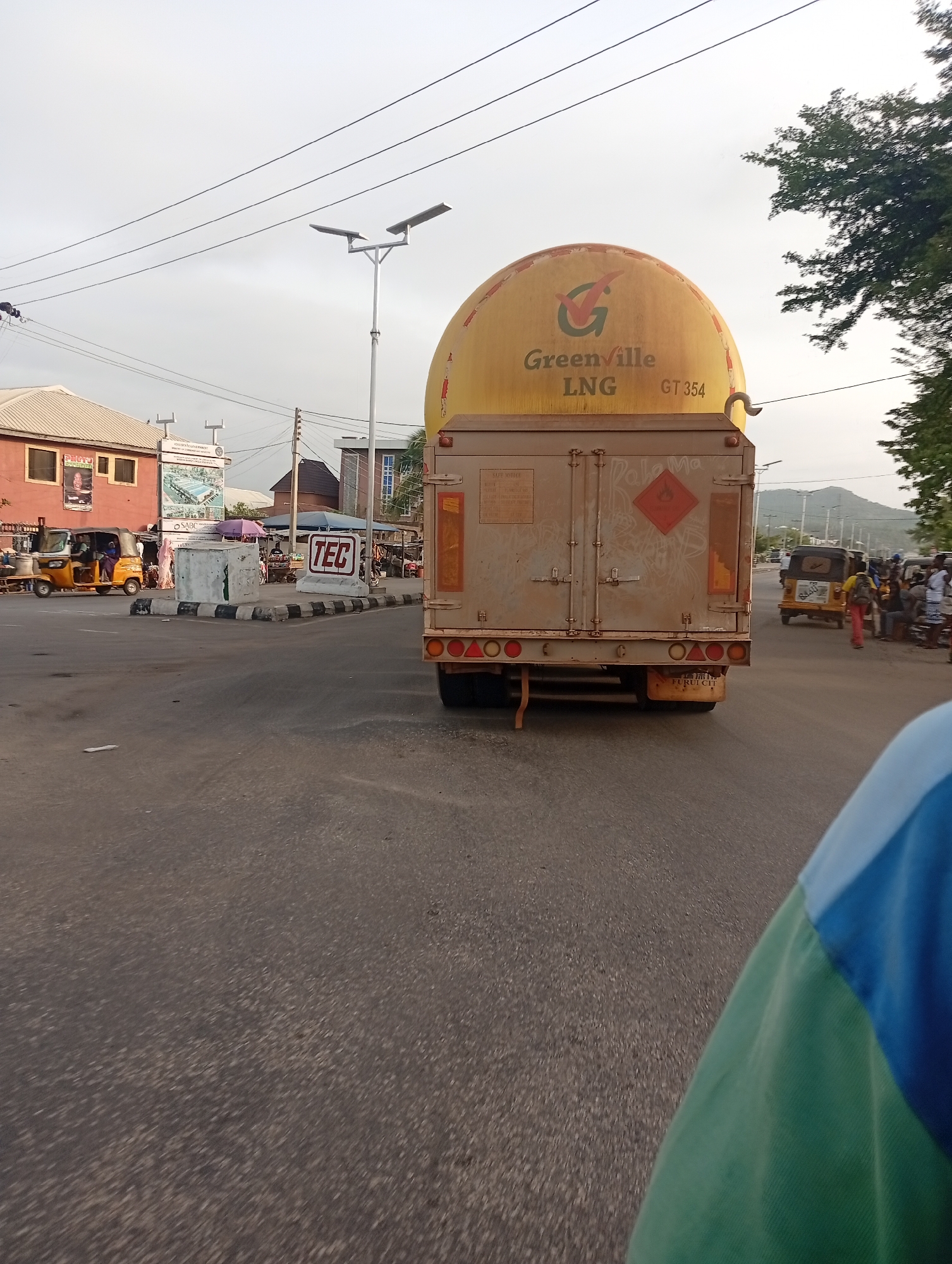 The image depicts a street scene in Adankolo, Nigeria, featuring a large, tanker truck in the foreground with "Greenville LNG" printed on its yellow spherical tank. The truck is stopped at a junction. To the left of the truck, there's a building with advertisement posters. Three-wheeled auto-rickshaws ("Keke Napep") are visible parked near the building. A "TEC" sign can be seen, indicating a technical or industrial presence. People stand around the junction. A mountain is visible in the background. In the immediate foreground on the lower right a person's arm in a blue and green shirt is visible. A modern streetlight is above the tanker. The road is paved and shows wear.