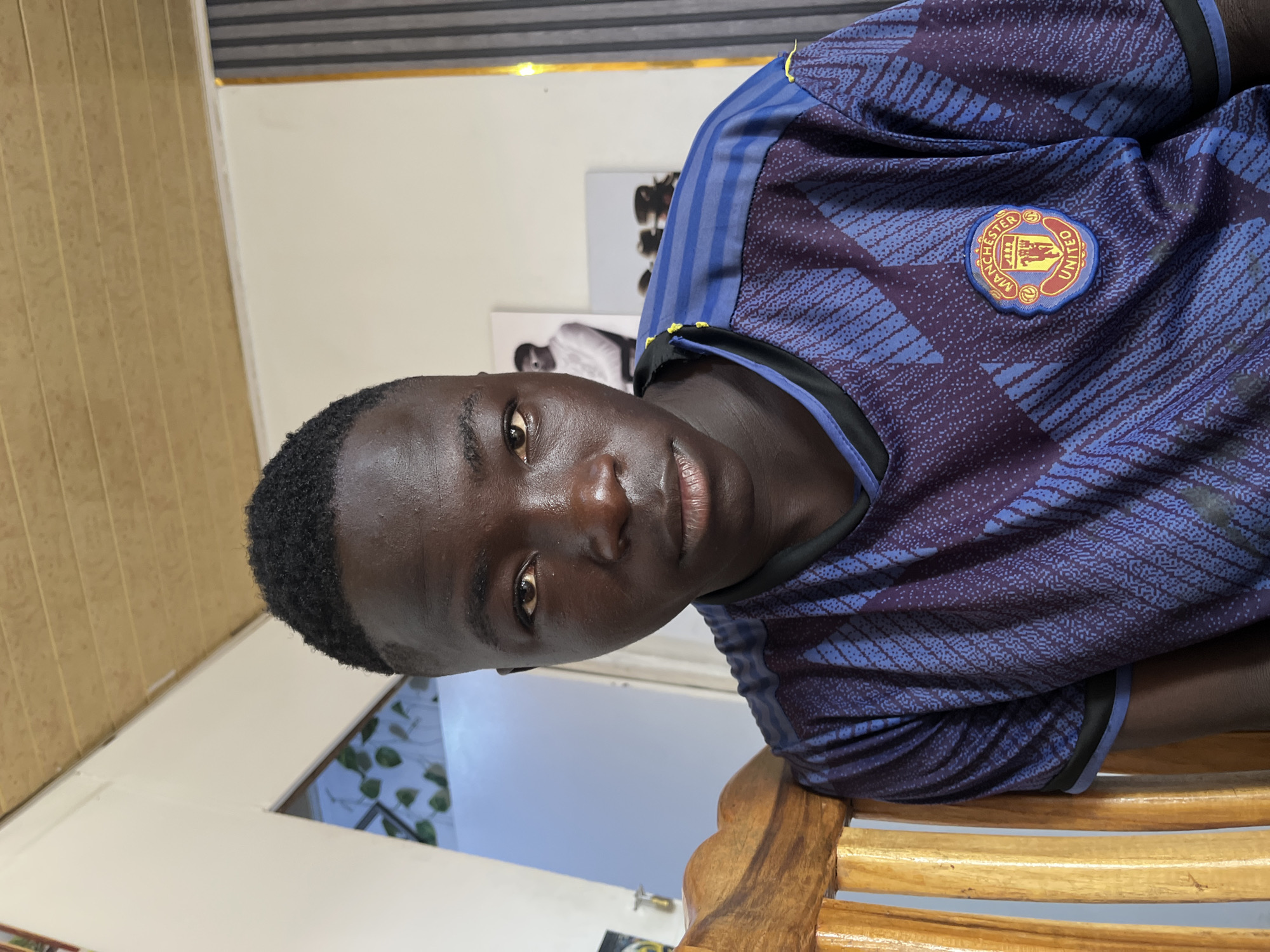 A medium-shot, slightly rotated image shows a dark-skinned young man wearing a blue Manchester United soccer jersey, sitting on a wooden chair. The background is a brightly lit room, with what appears to be window louvers and artwork on the wall. There is no visible evidence to definitively confirm the location as Jos, Nigeria.