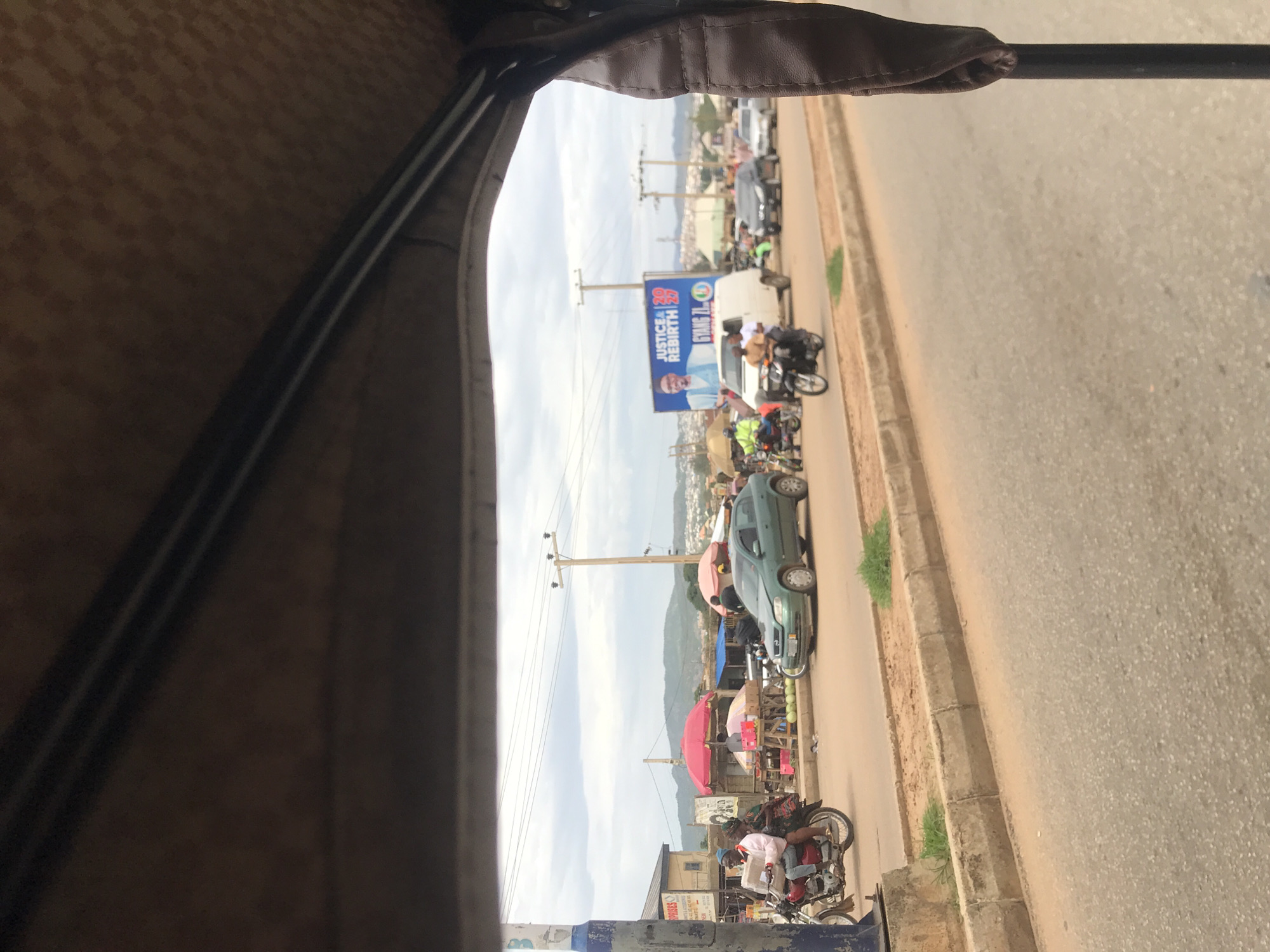 The image presents a street-level view from the interior of a vehicle, likely a tricycle, as indicated by the patterned brown fabric and metal frame in the foreground. The scene depicts an active urban thoroughfare in Jos, Plateau State, Nigeria.
On the left side of the road, a red motorcycle with two male occupants is partially visible, positioned near a blue and white utility pole. The passenger is carrying a package. Behind them, market stalls with red and orange umbrellas display various goods, including watermelons, beside a building marked "ENTERPRISES."
Traffic flows on a paved road with a low concrete median strip. Vehicles observed include a green hatchback car with a slightly damaged front bumper, a white panel van, and multiple motorcycles with single riders. In the mid-ground, a prominent blue billboard features a man in a light blue suit and glasses, with the text "JUSTICE & REBIRTH 2027" and "GYANG ZL." This political advertisement serves as a key indicator of the location.
Numerous wooden utility poles with overhead power lines line both sides of the street. The sky is overcast with grey clouds. In the background, distant hills are visible, with scattered residential structures on their slopes, characteristic of the topography around Jos.
