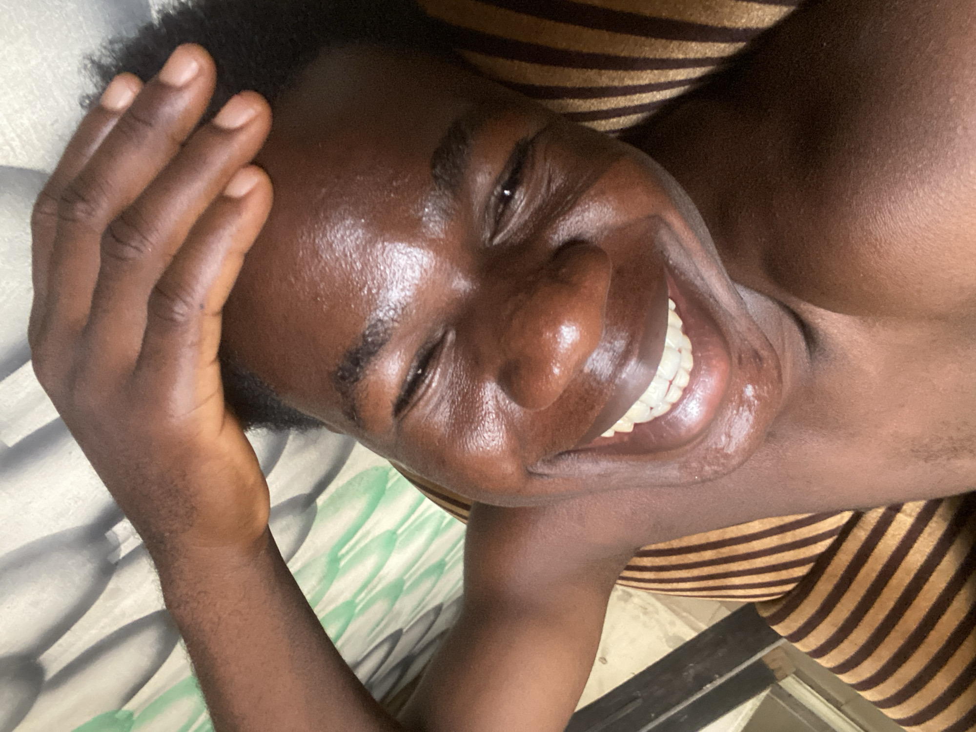 A close-up, tilted shot of a dark-skinned individual, seemingly lying down, with their right hand partially obscuring their forehead and hair. The person is smiling widely, revealing a full set of teeth. The skin appears shiny, possibly due to sweat or oil. The background includes a striped surface, likely a piece of furniture or bedding, and a light-colored surface with indistinct patterns. There is no information in the image to locate this scene in Mararaba, Nigeria.